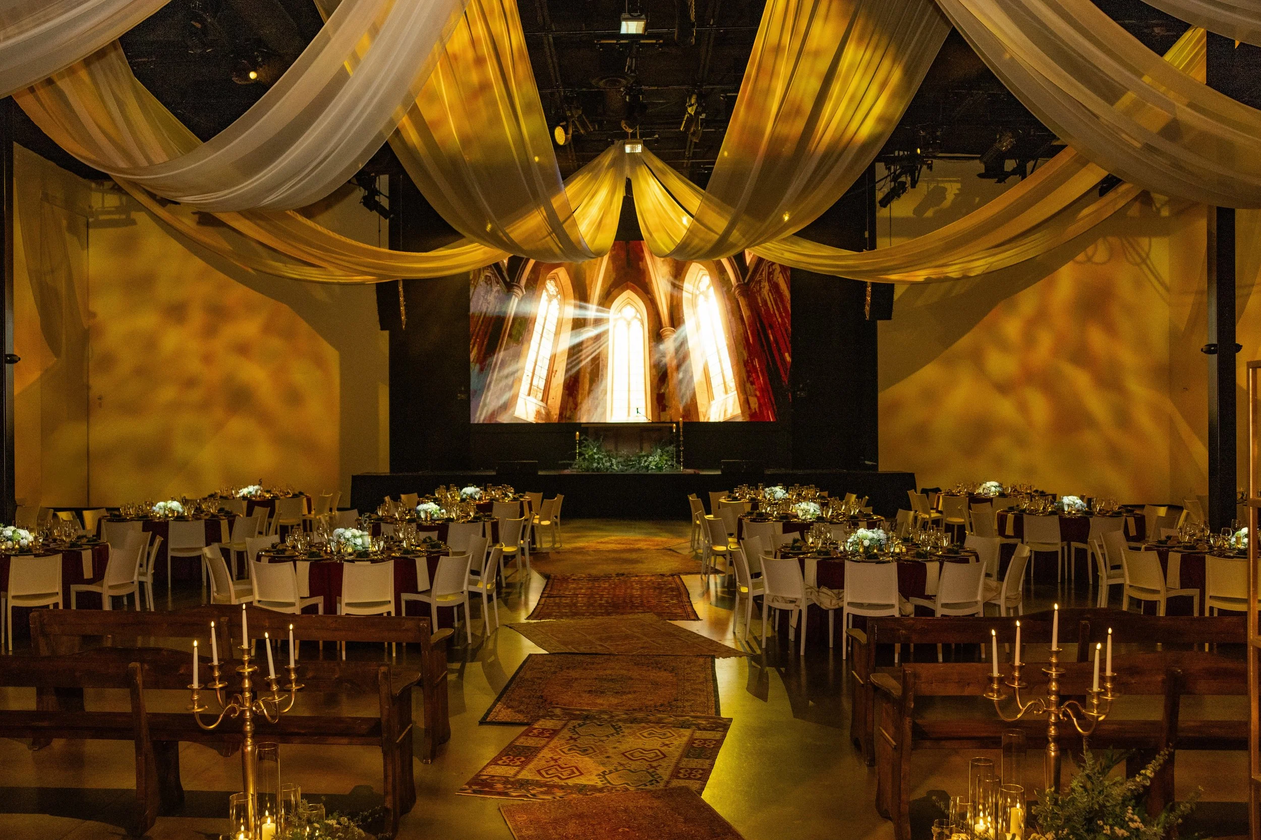 A banquet hall decorated for a special event with round tables covered in white tablecloths, floral centerpieces, gold candles, and gold chairs. The high ceiling features flowing white and gold drapes. There is a large projection screen showing an im