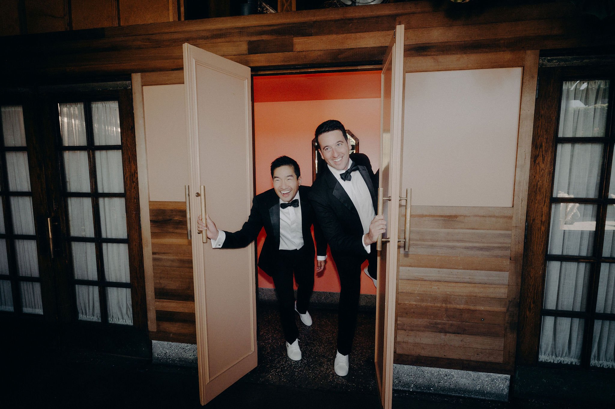 Two men in tuxedos with bow ties are smiling and leaning through a small bamboo door in a wooden wall, inside a room with pink and orange walls.
Cliff + Warren wedding. Smog Shoppe, Culver City