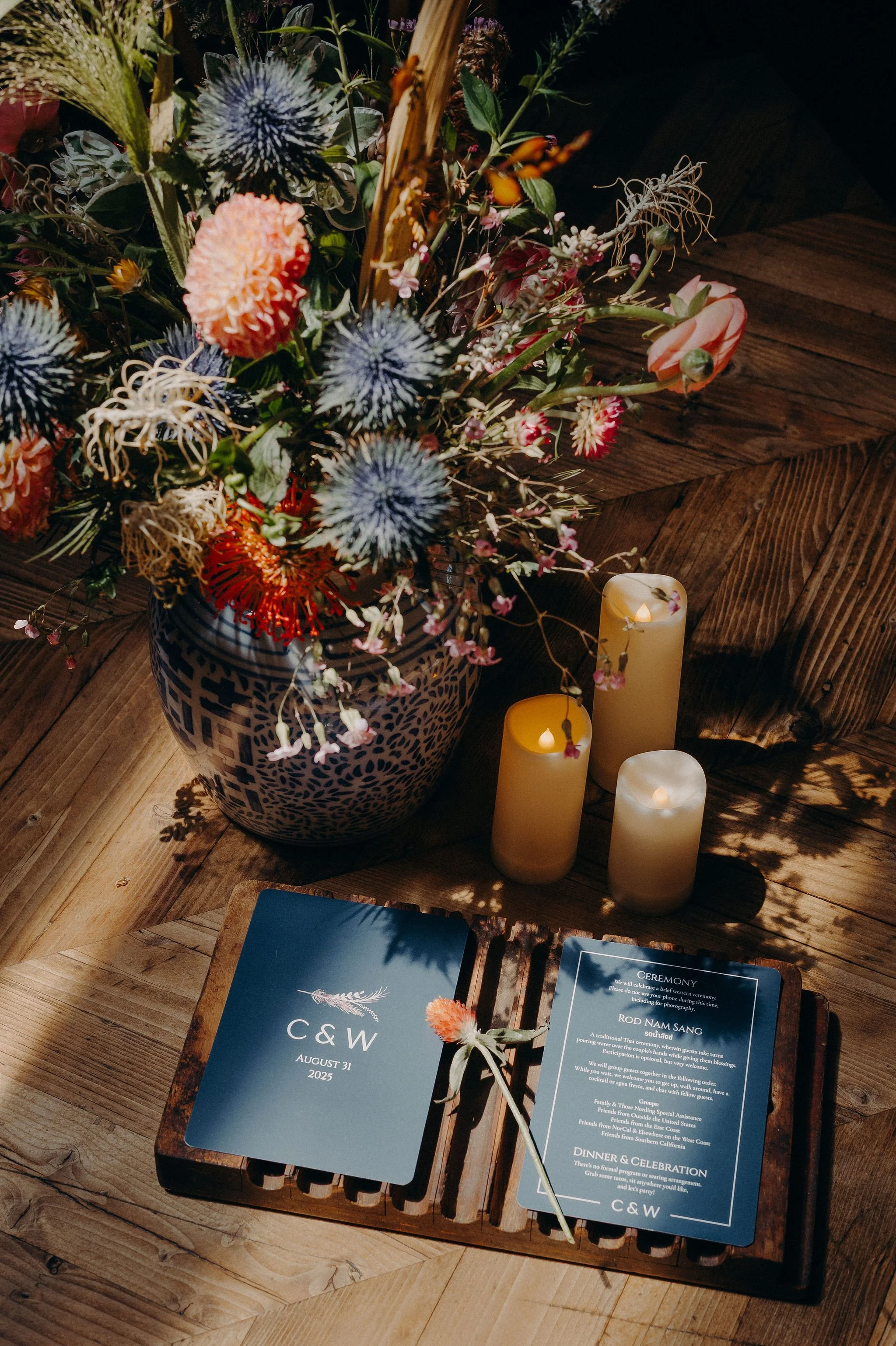 A wooden table with a blue and white patterned vase containing various colorful flowers, surrounded by three LED candles and a wedding program with the initials C&W and the date August 31, 2025.
Cliff + Warren wedding. Smog Shoppe, Culver City