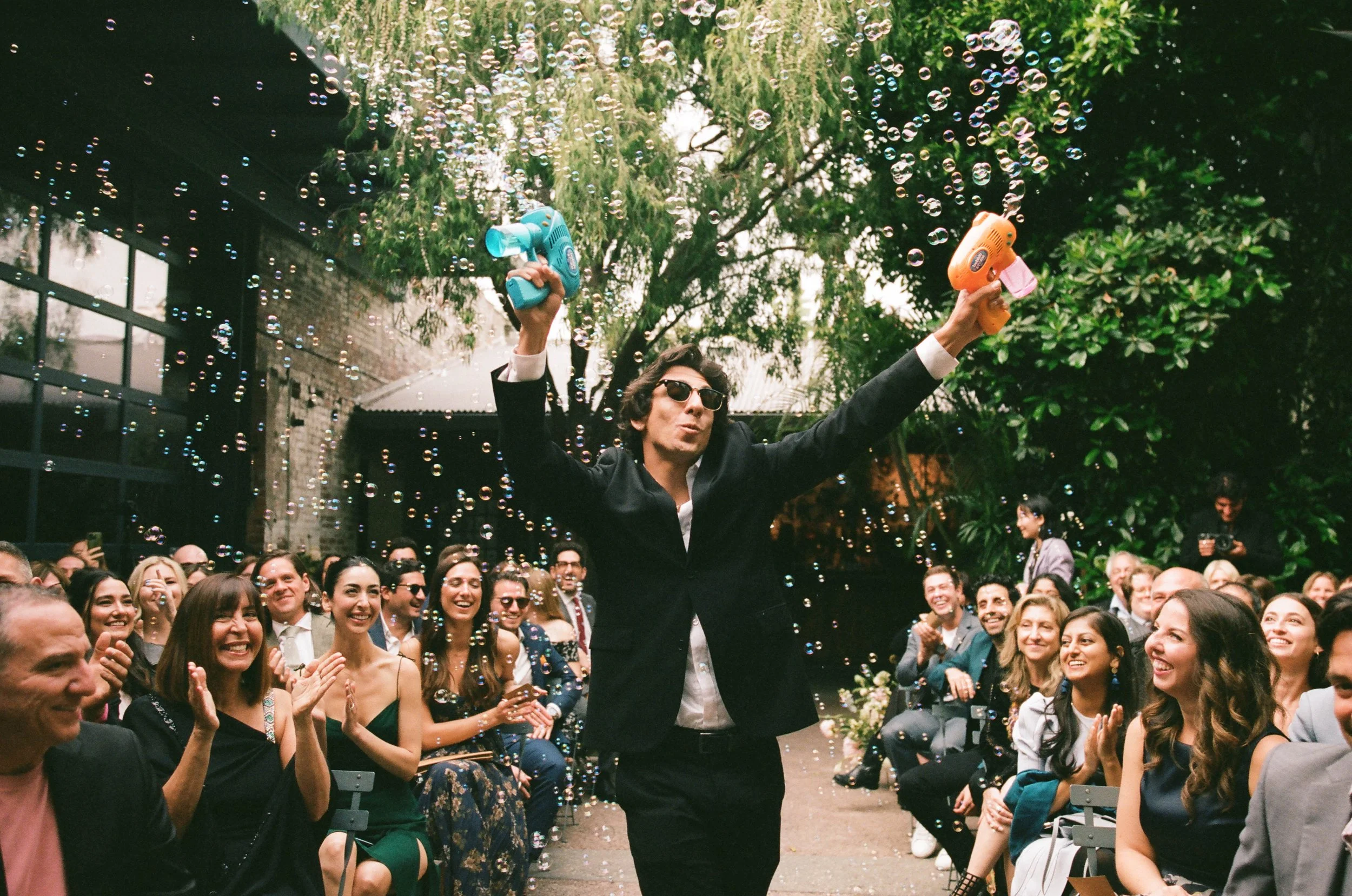 A man in a suit and sunglasses holding two bubble machines, standing in front of a large crowd outdoors, surrounded by bubbly soap and greenery, with people smiling, clapping, and enjoying the event.
Carolyn + Ben Wedding, Millwick DTLA