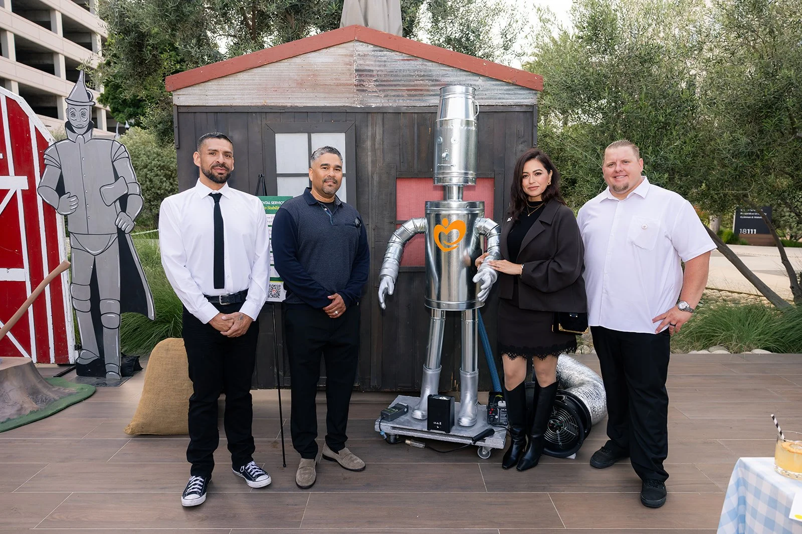 Four people standing outdoors in front of a small wooden shed, with a large metallic robot sculpture in the center. The group includes three men and one woman, all dressed in business casual or casual attire. There are trees and a parking structure i