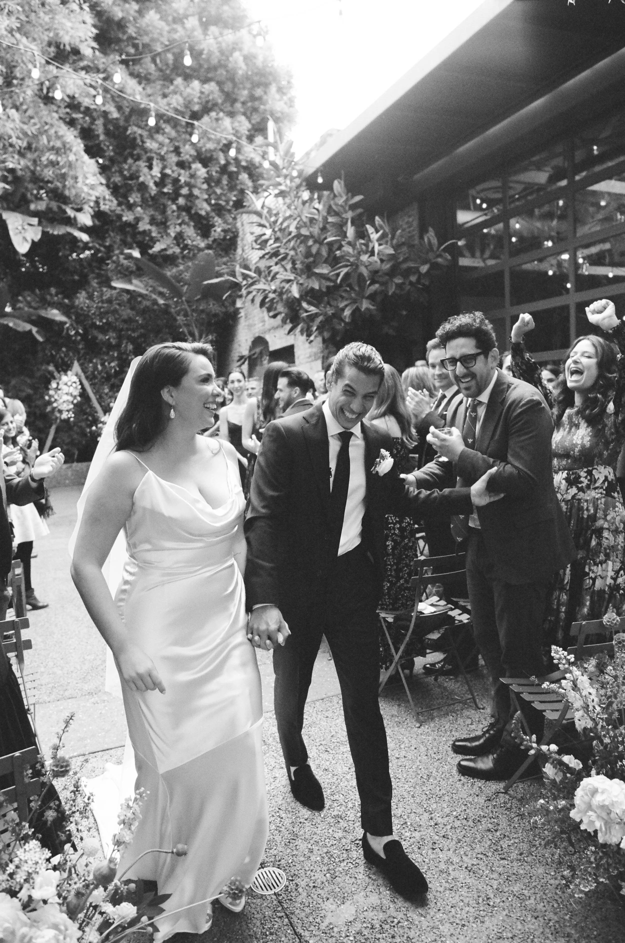 Black and white photo of a wedding celebration outdoors. The bride and groom are holding hands and smiling, surrounded by friends and family who are cheering. The bride is wearing a sleeveless gown and veil, and the groom is in a dark suit. Decoratio