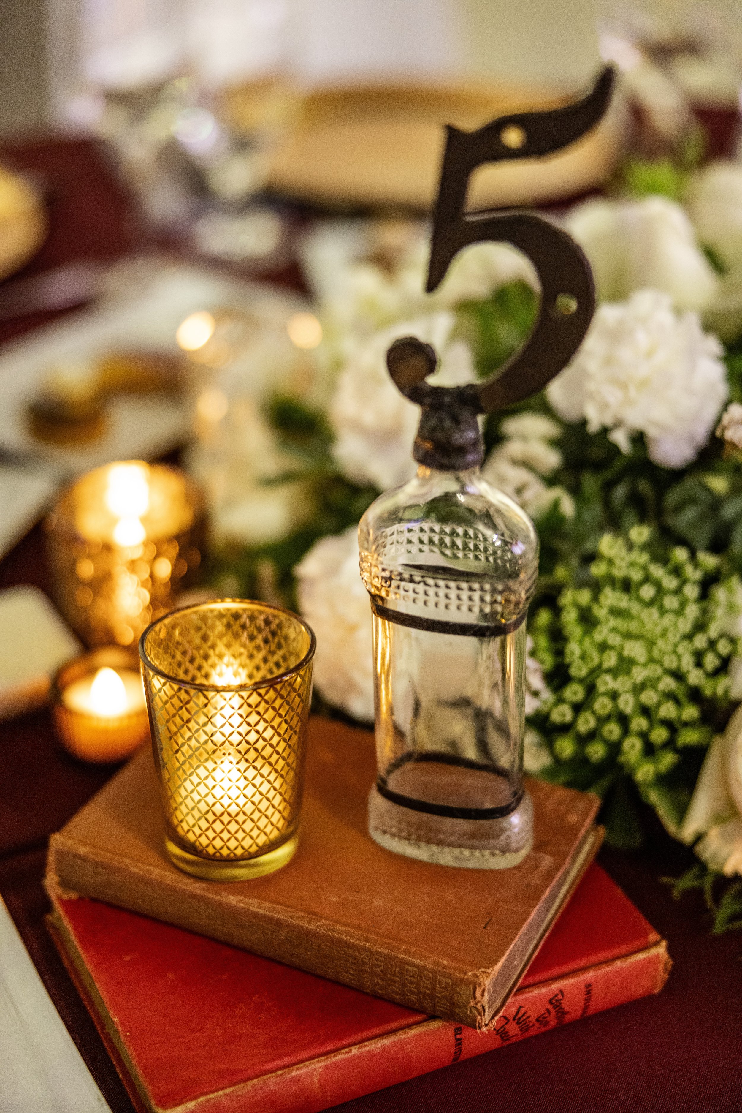 Decorative scene with a gold candle holder, a glass bottle, and a black number five candle, all resting on a vintage hardcover book, with flowers and soft lighting in the background.