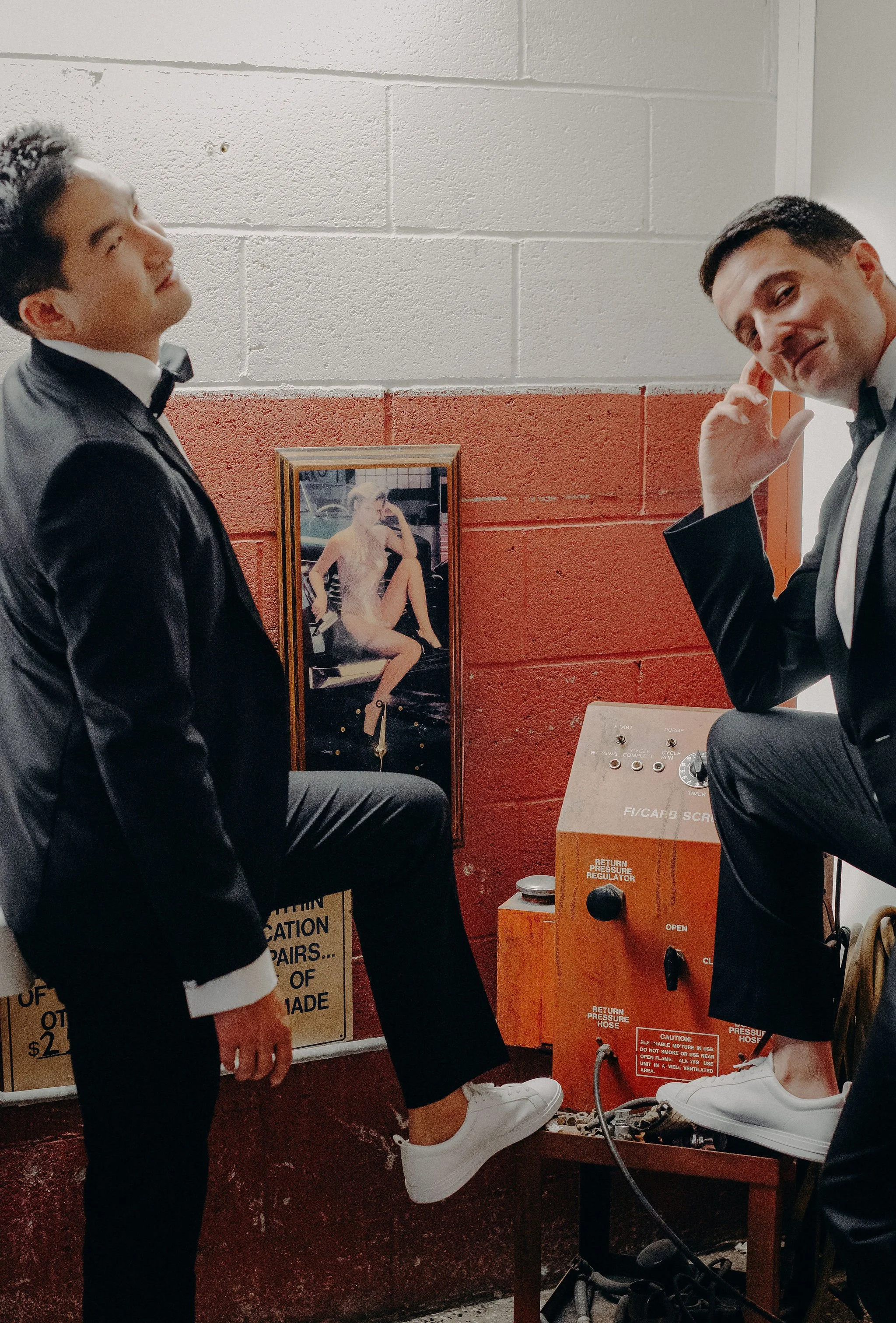 Two men in tuxedos beside a carpet cleaning machine with a framed picture of a woman. One man is standing, the other is sitting with his foot on the machine, and they are talking.
Cliff + Warren wedding. Smog Shoppe, Culver City
