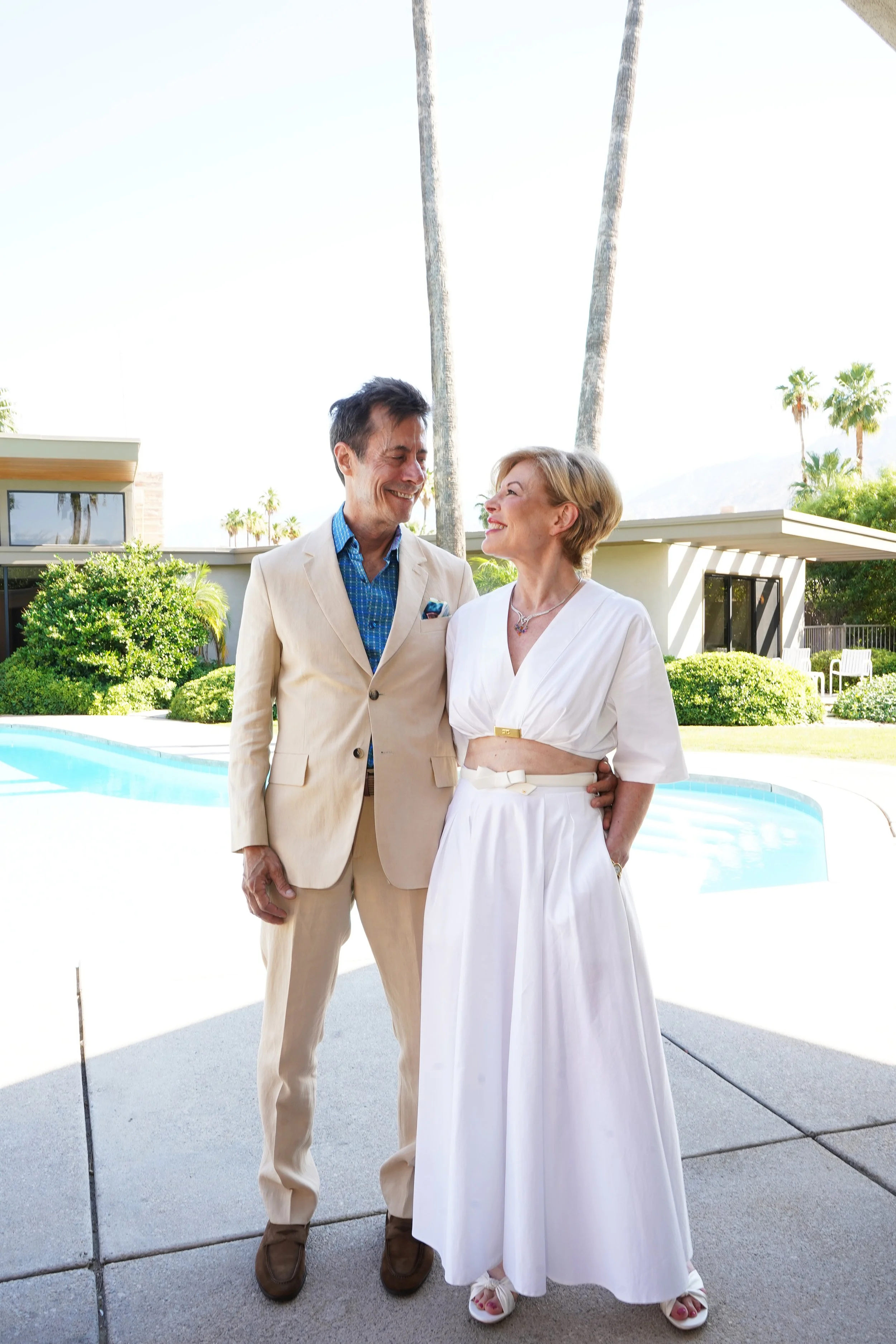 A man and woman smiling and standing outdoors near a swimming pool, with palm trees and mid-century modern house in the background.