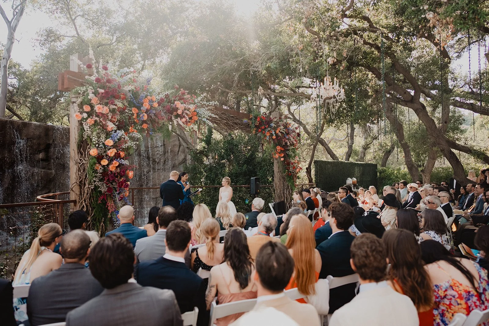 Outdoor wedding ceremony under trees with hanging chandeliers, large floral arch, and waterfall backdrop, with seated guests watching the couple exchange vows.