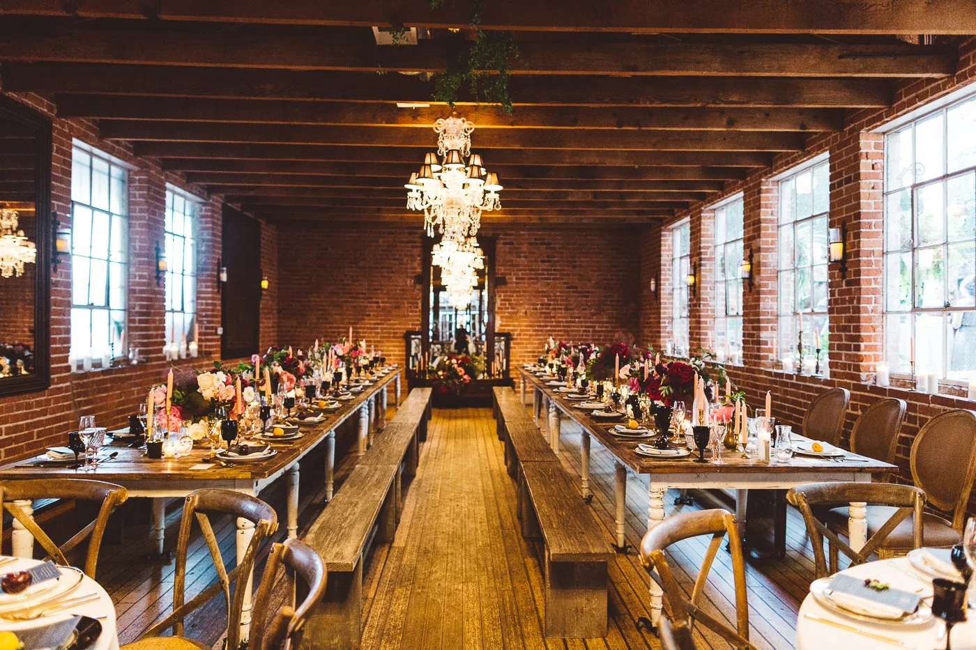 Elegant indoor banquet hall with exposed brick walls, large windows, chandeliers, and long tables decorated with flowers, candles, and table settings.