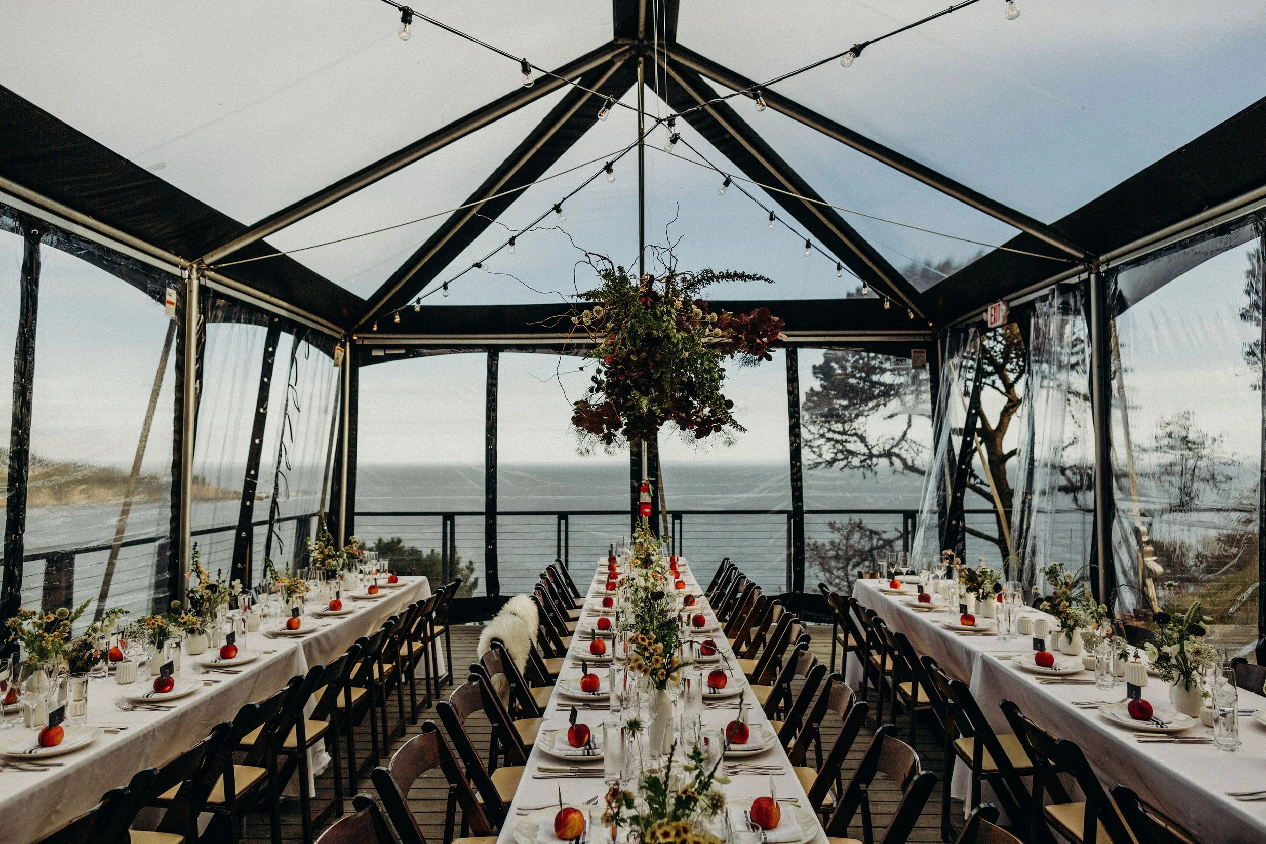 Long dining tables with white tablecloths decorated with flowers and small pumpkins, set up for a meal inside a transparent tent with string lights hanging from above, overlooking a ocean view modern decor. Amy + Dave, Timber Cove 