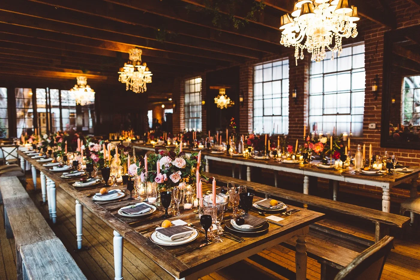 A long wooden banquet table set for a formal event with flower arrangements, candles, and tableware in a rustic-style room with brick walls, large windows, and chandeliers hanging from the ceiling.