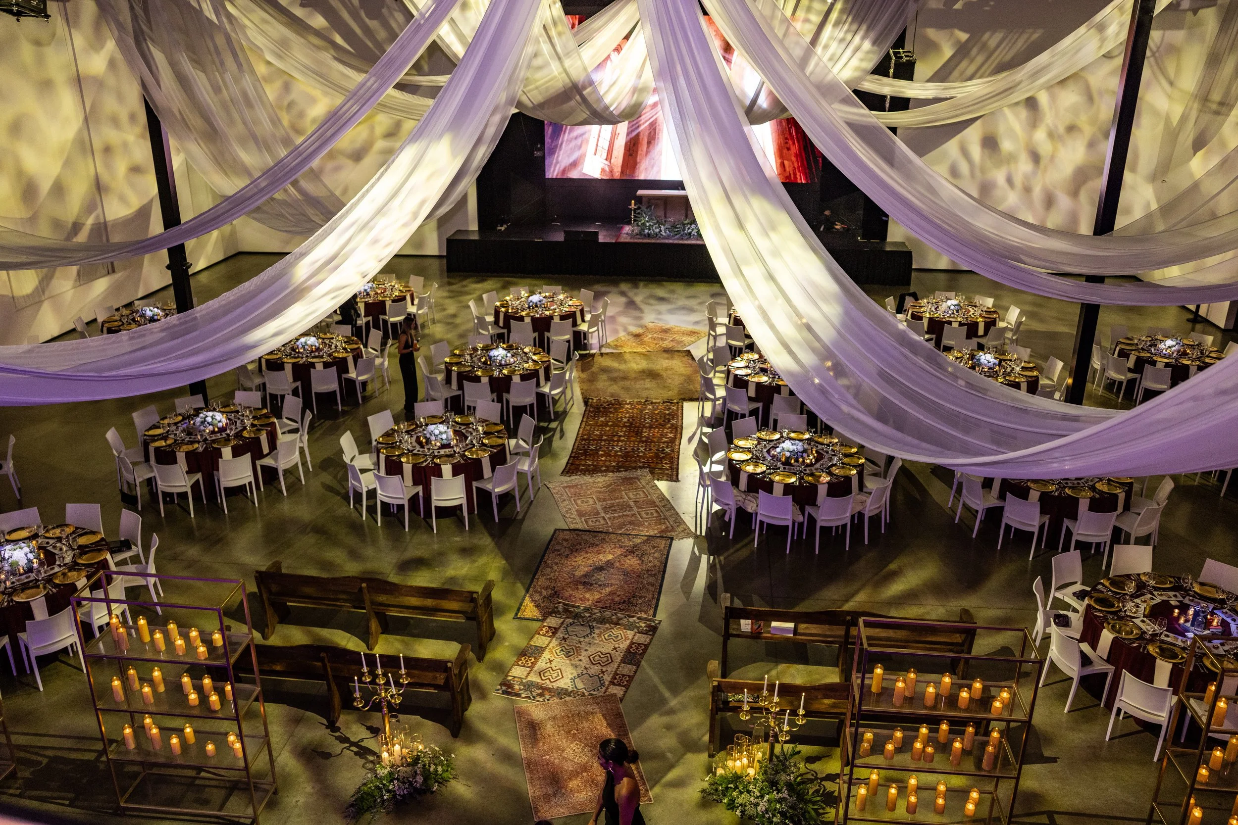 Elegant event space decorated with flowing white fabric drapes overhead, arranged around round tables with gold and white settings, candles, and floral centerpieces. A stage with a large screen is at the front, and multiple patterned rugs are laid across the floor, creating a warm, inviting atmosphere for a celebration or gathering.