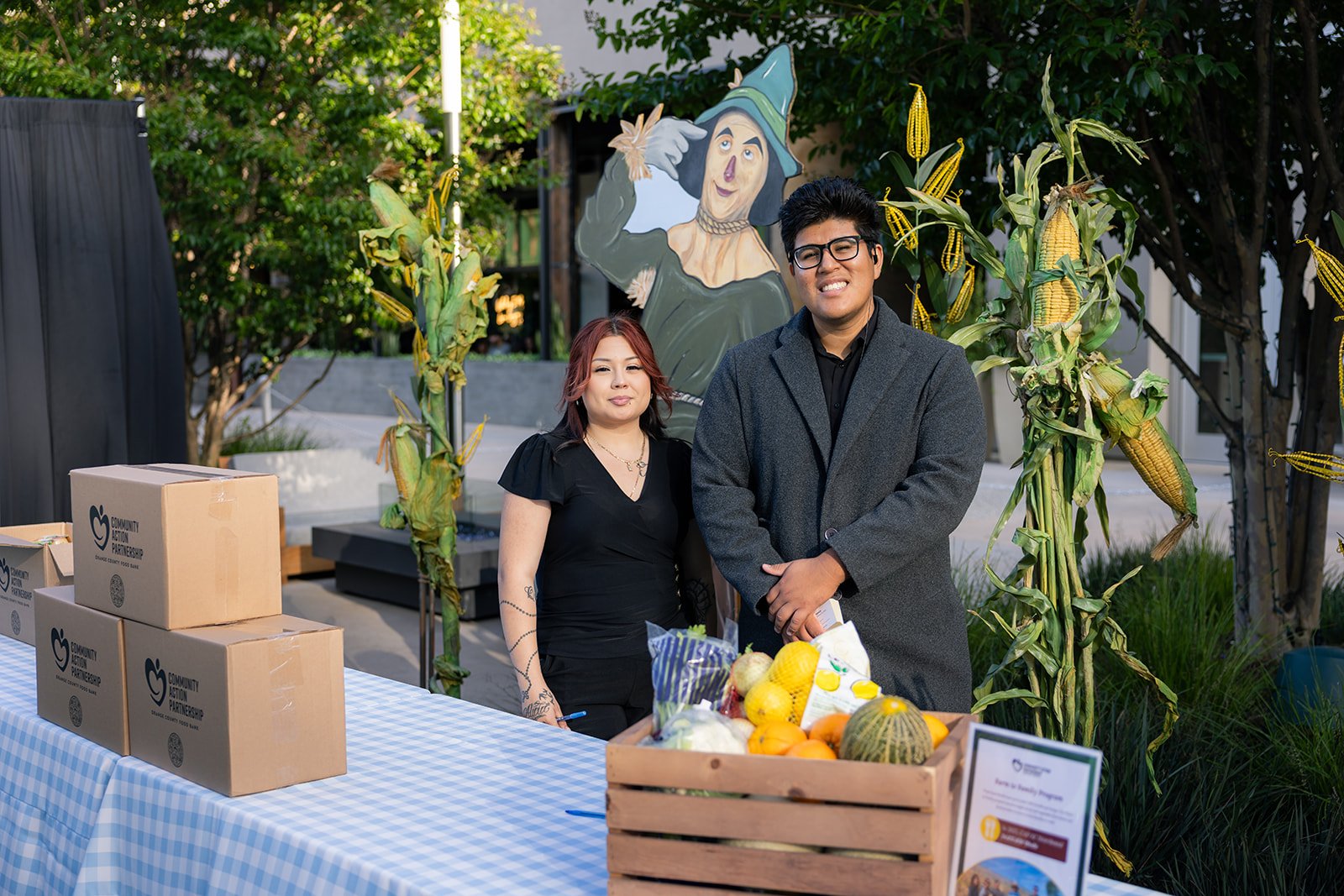 Two people standing behind a table with boxes and produce at an outdoor event, with a large animated witch figure in the background and corn stalks decorated around them.