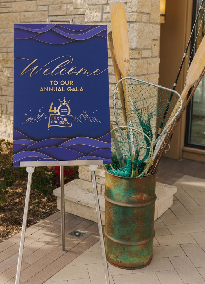 A blue sign with gold text welcoming guests to an annual gala celebrating 40 years for the Children, with mountain and star illustrations. Next to the sign is a rusty, greenish barrel holding a fishing net and wooden oars, placed outside near a brick wall.