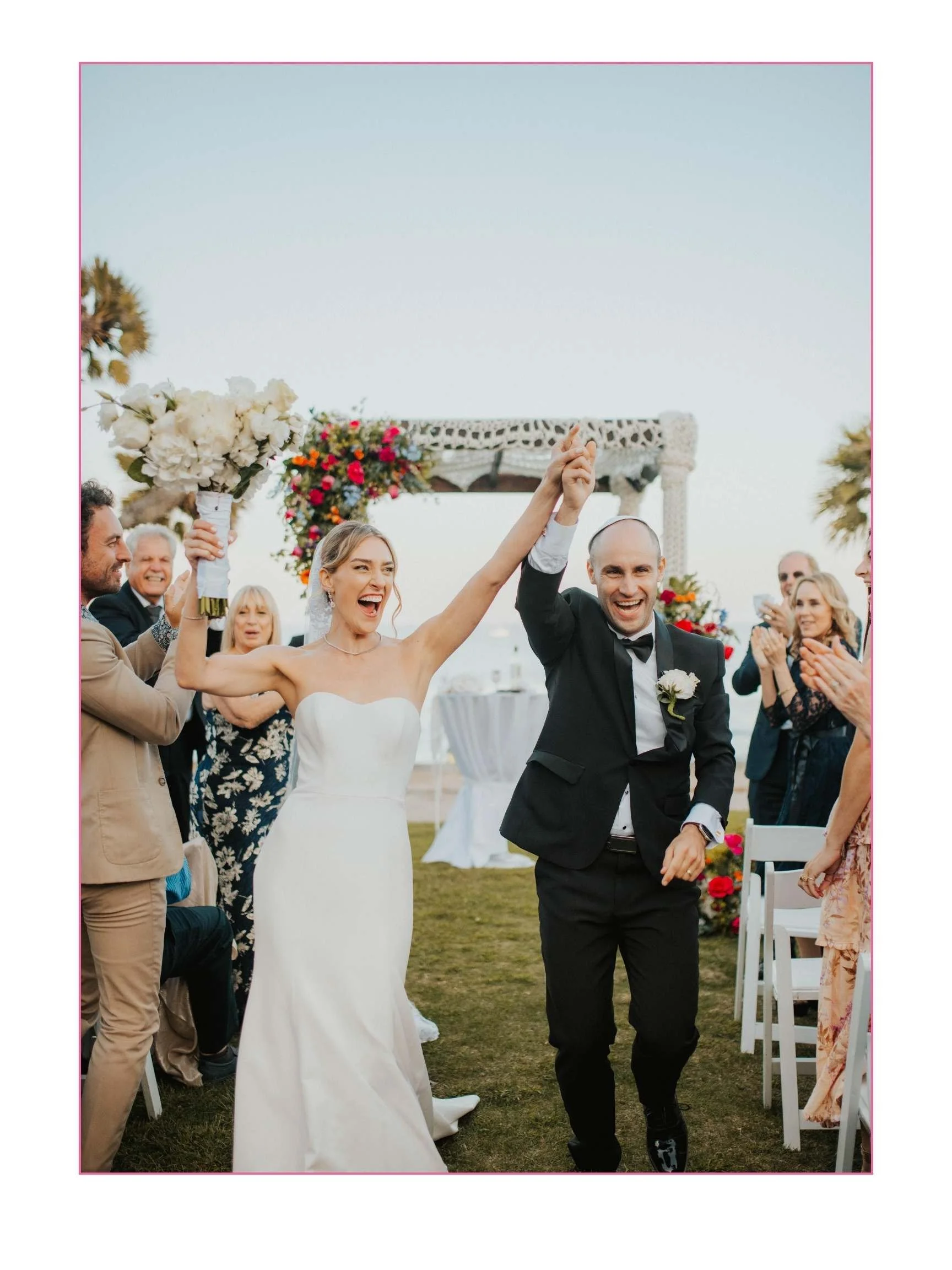 A bride and groom celebrating their wedding, holding hands and smiling amid guests outdoors during sunset, with floral arrangements and a wedding arch in the background.