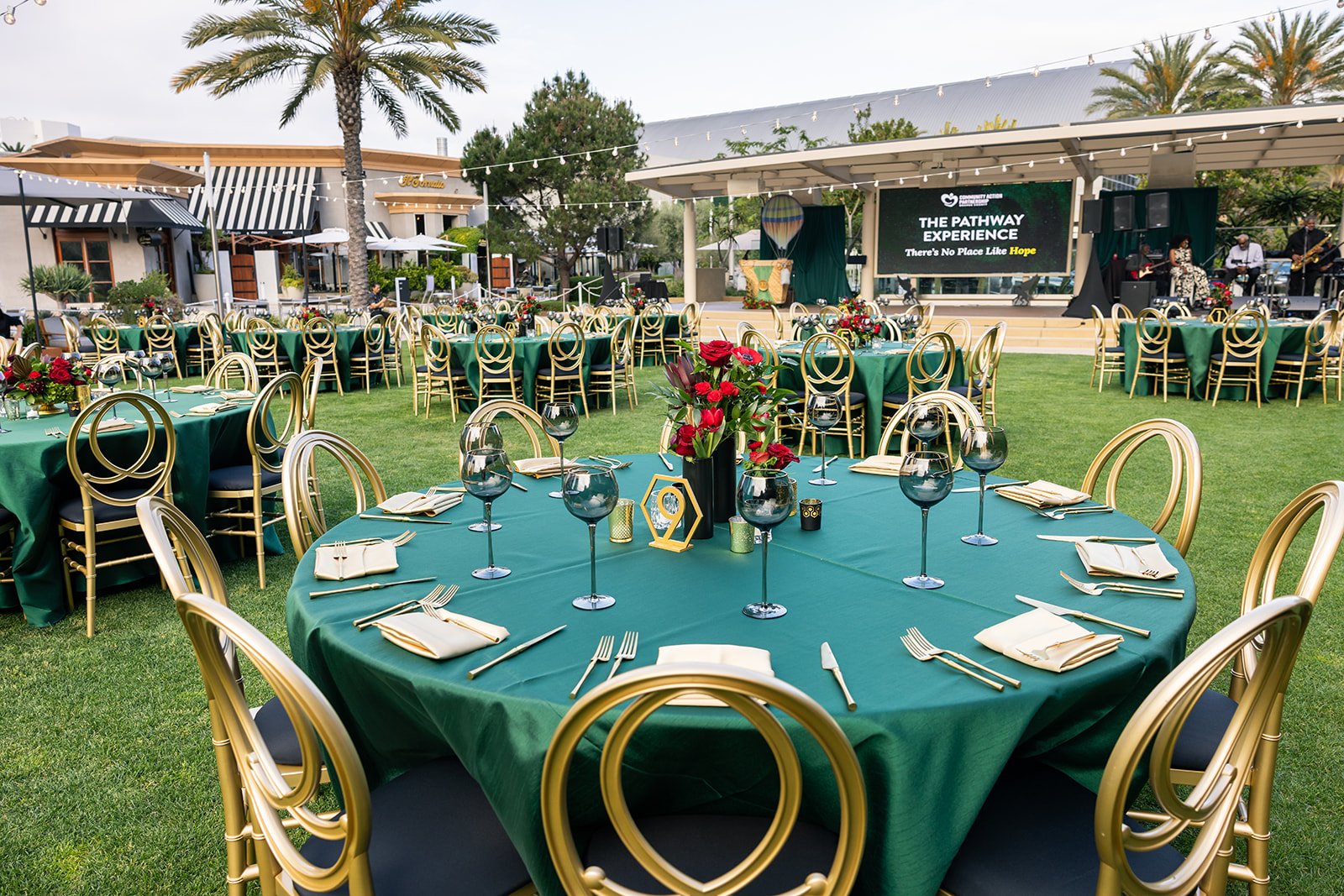 Outdoor event setup with round tables decorated with green tablecloths, gold chairs, floral centerpieces, and place settings. A stage with a large screen displaying 'The Pathway Experience' and live band nearby, with palm trees and buildings in the b