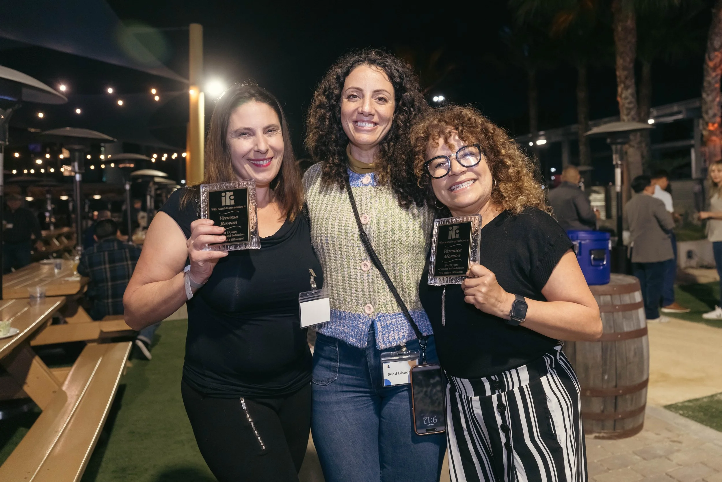 Three women smiling and holding awards at an outdoor event during night time.