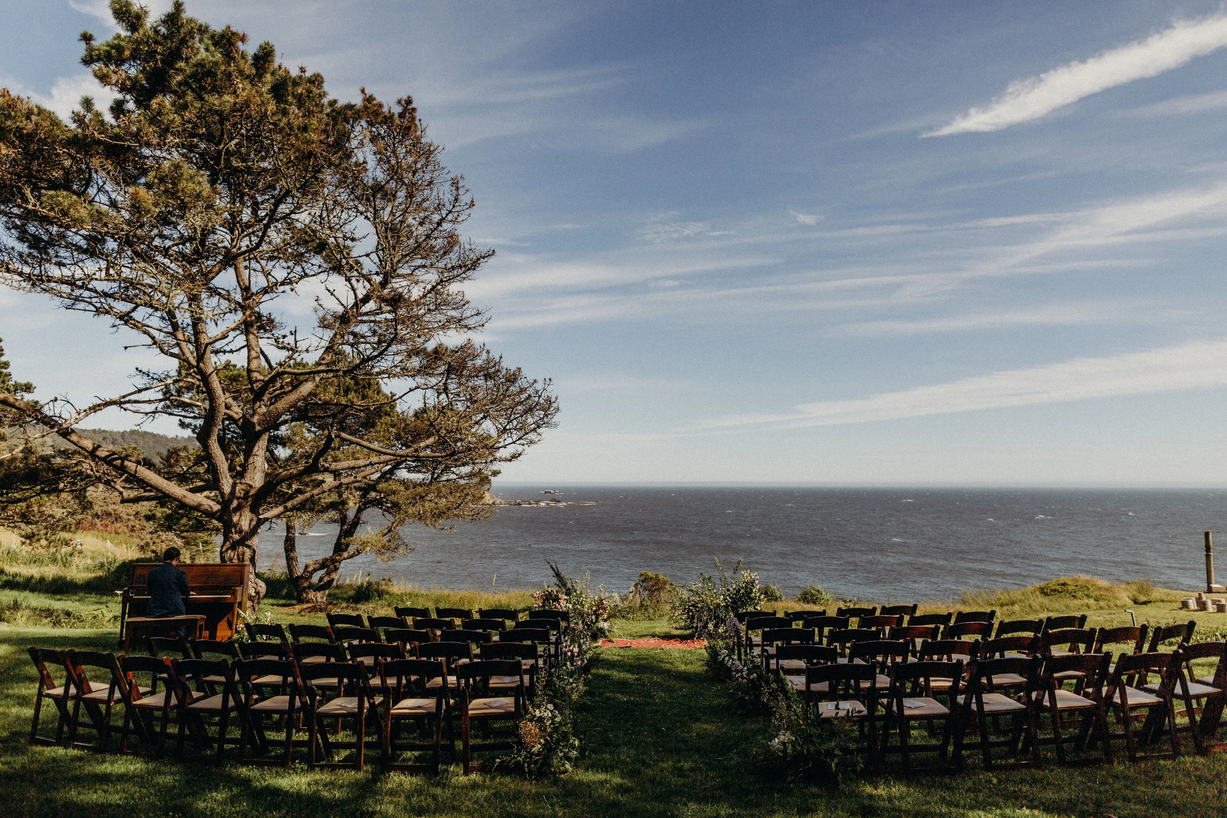 Outdoor event setup at a grassy seaside location with chairs arranged in rows, a large leafy tree, a person playing piano, and ocean in the background under a partly cloudy sky. Amy + Dave, Timber Cove 