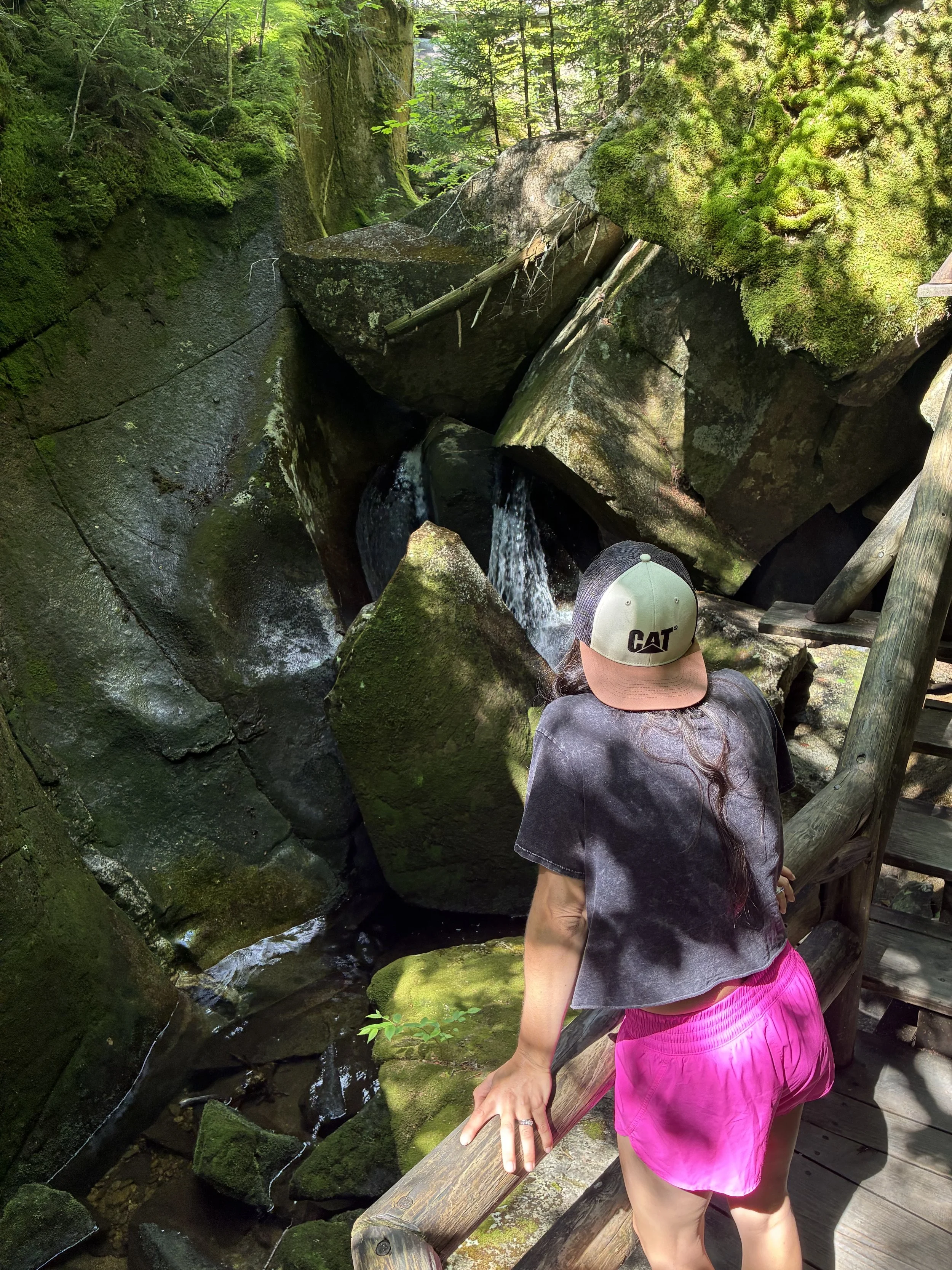 A person with long dark hair, wearing a beige and black Cat baseball cap, a black tie-dye t-shirt, and pink shorts, standing on a wooden bridge near a moss-covered rocky stream in a forest.