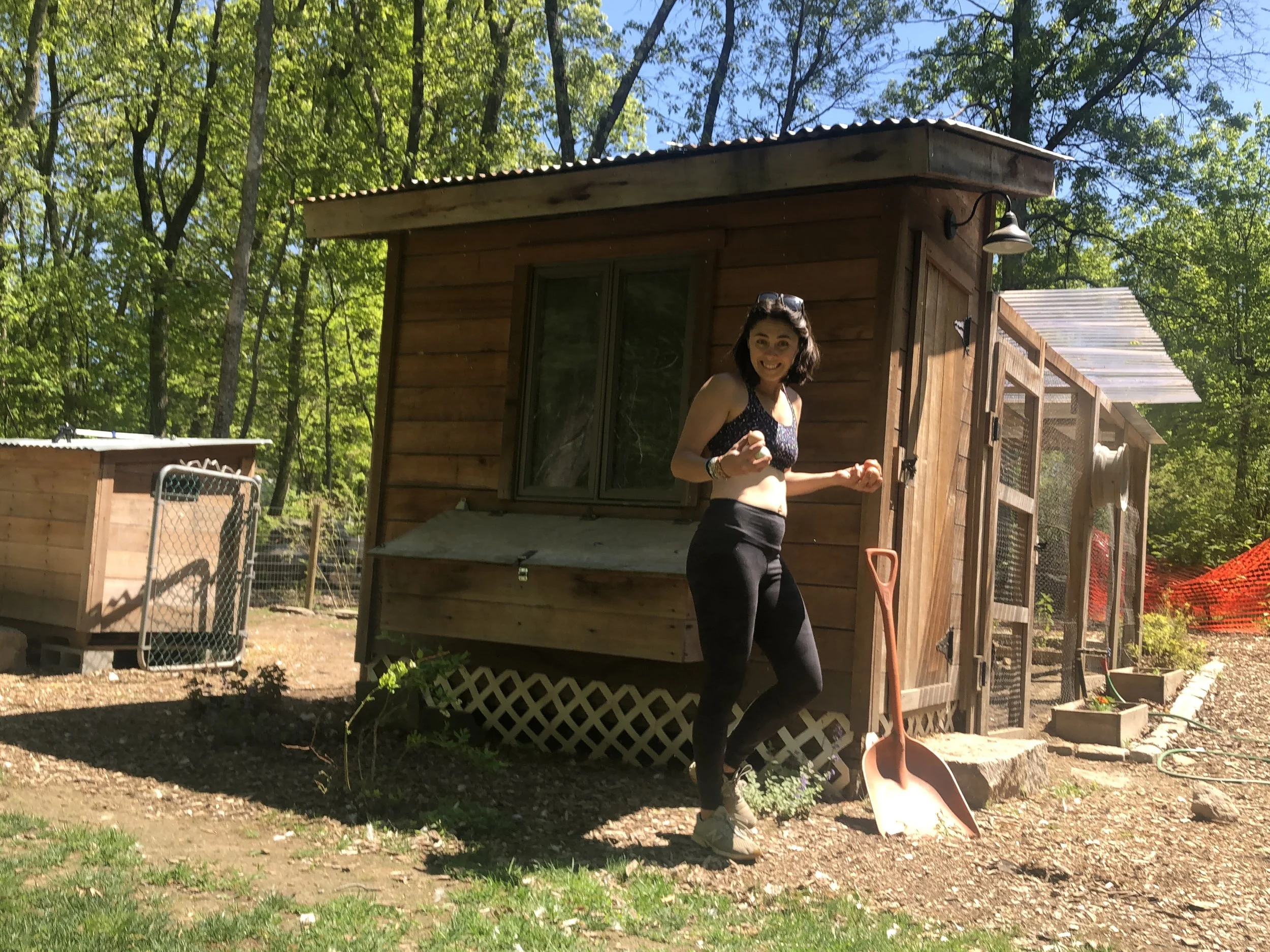 A woman standing outside near a wooden shed with trees and blue sky in the background. She is smiling, wearing a black top, and has sunglasses on her head, with a shovel leaning against the shed.
