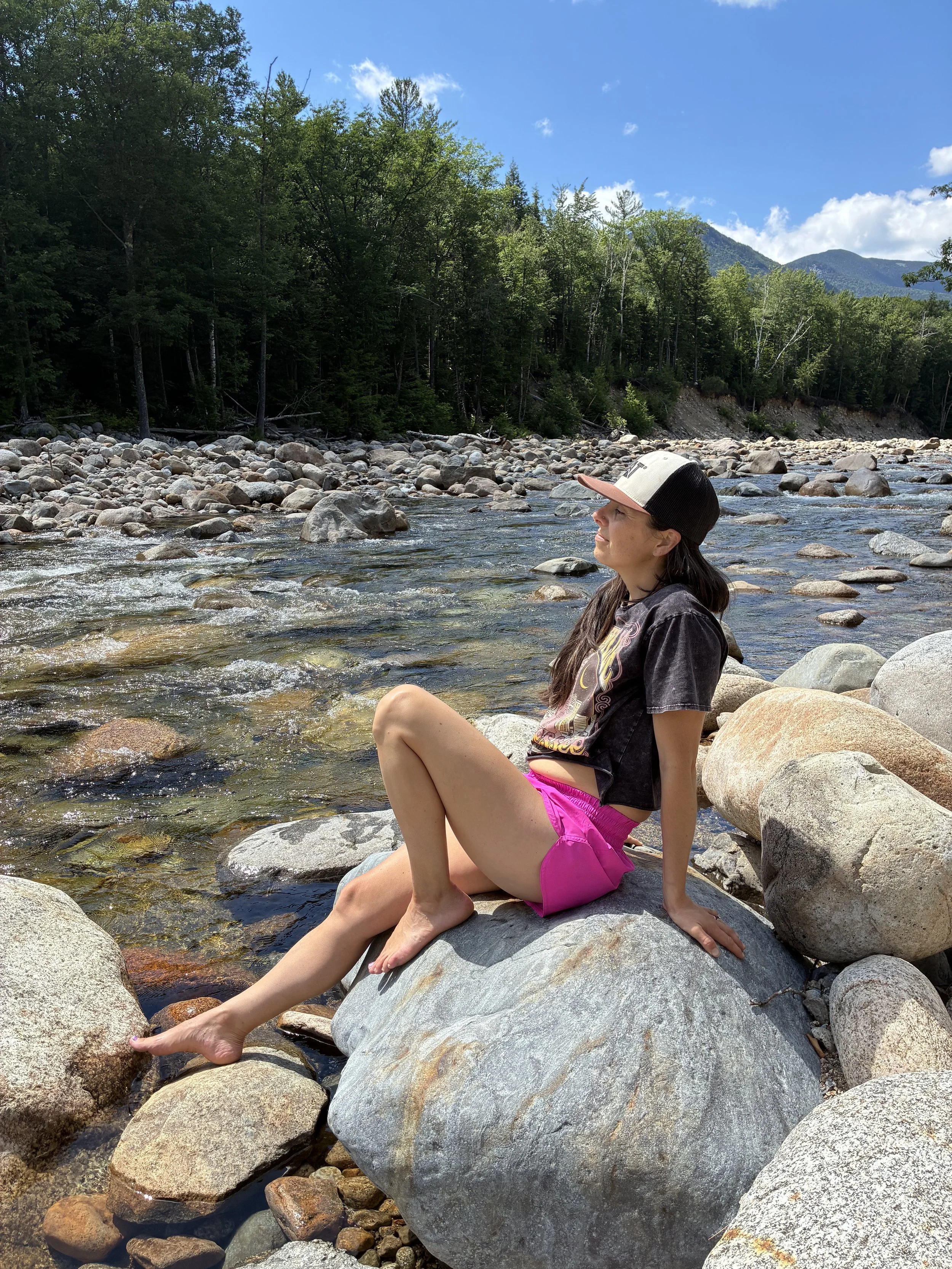 A young woman sits on a large rock by a shallow mountain stream, wearing a cap, a black t-shirt, and pink shorts, with her eyes closed and face tilted upward. The scene is outdoors with a backdrop of green trees and mountains under a partly cloudy sky.