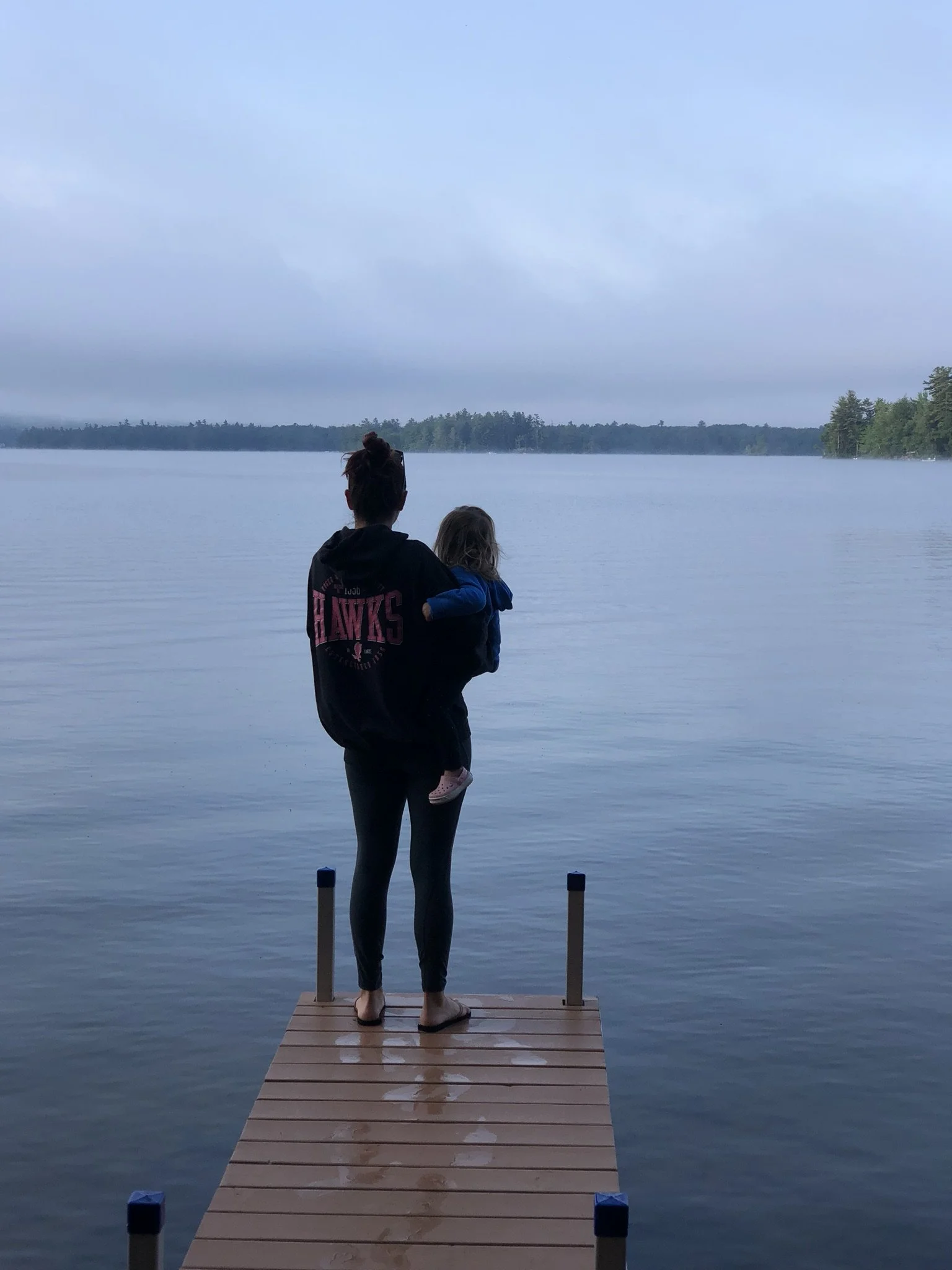A woman holding a young girl standing on a wooden dock at the edge of a calm lake with trees in the distance, during early morning or evening with overcast sky.