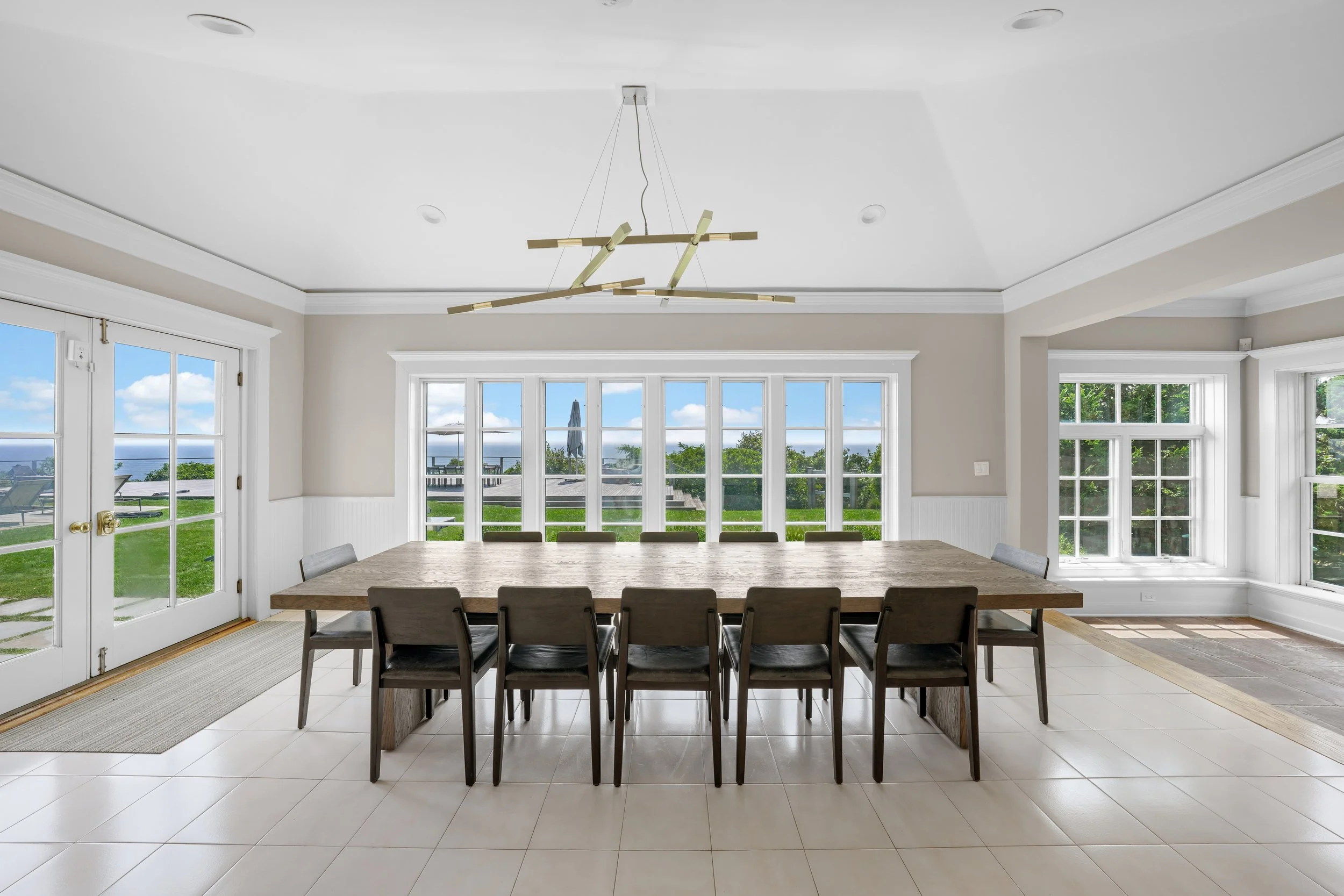 Empty dining room with large windows showing outdoor greenery and ocean view, beige walls, modern ceiling light fixture, wooden dining table, and ten black chairs.