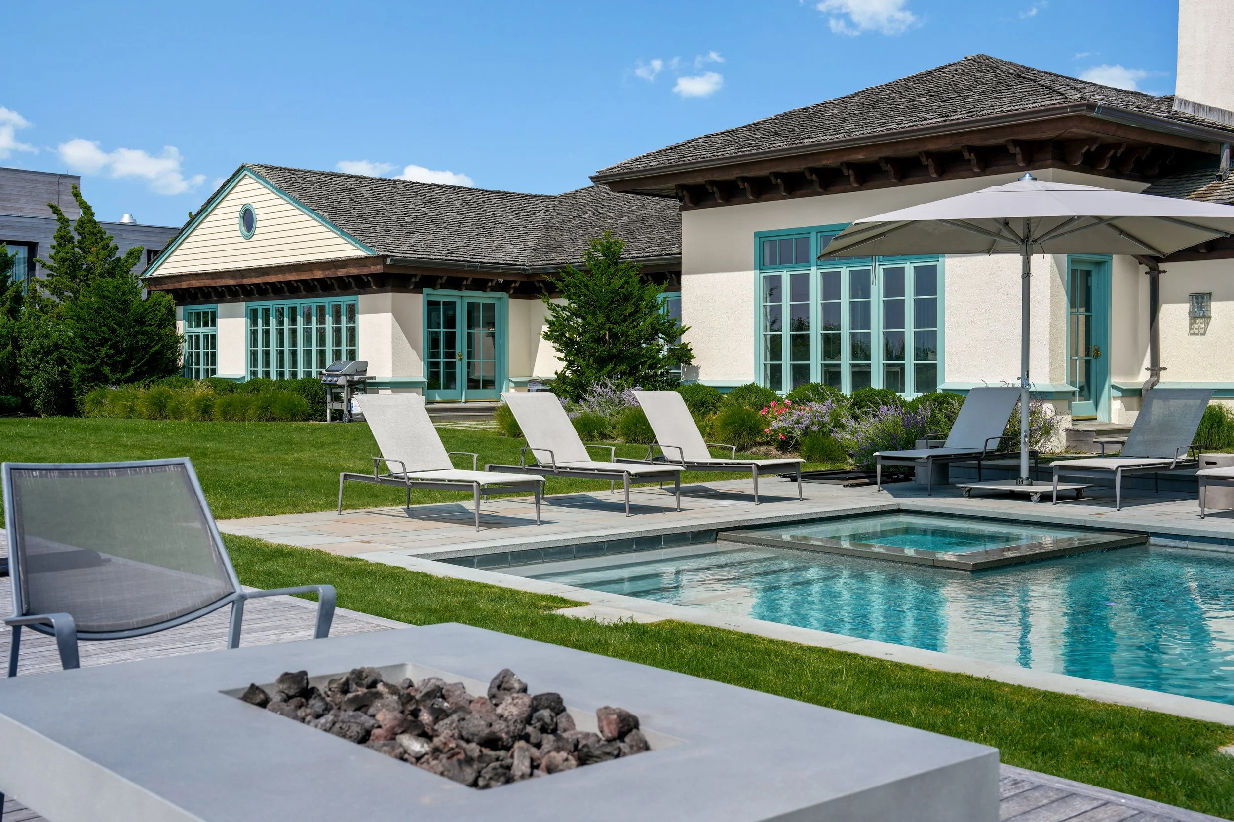 Luxury backyard with a swimming pool, lounge chairs, a white umbrella, and a house with large windows and a patio area under clear blue skies.