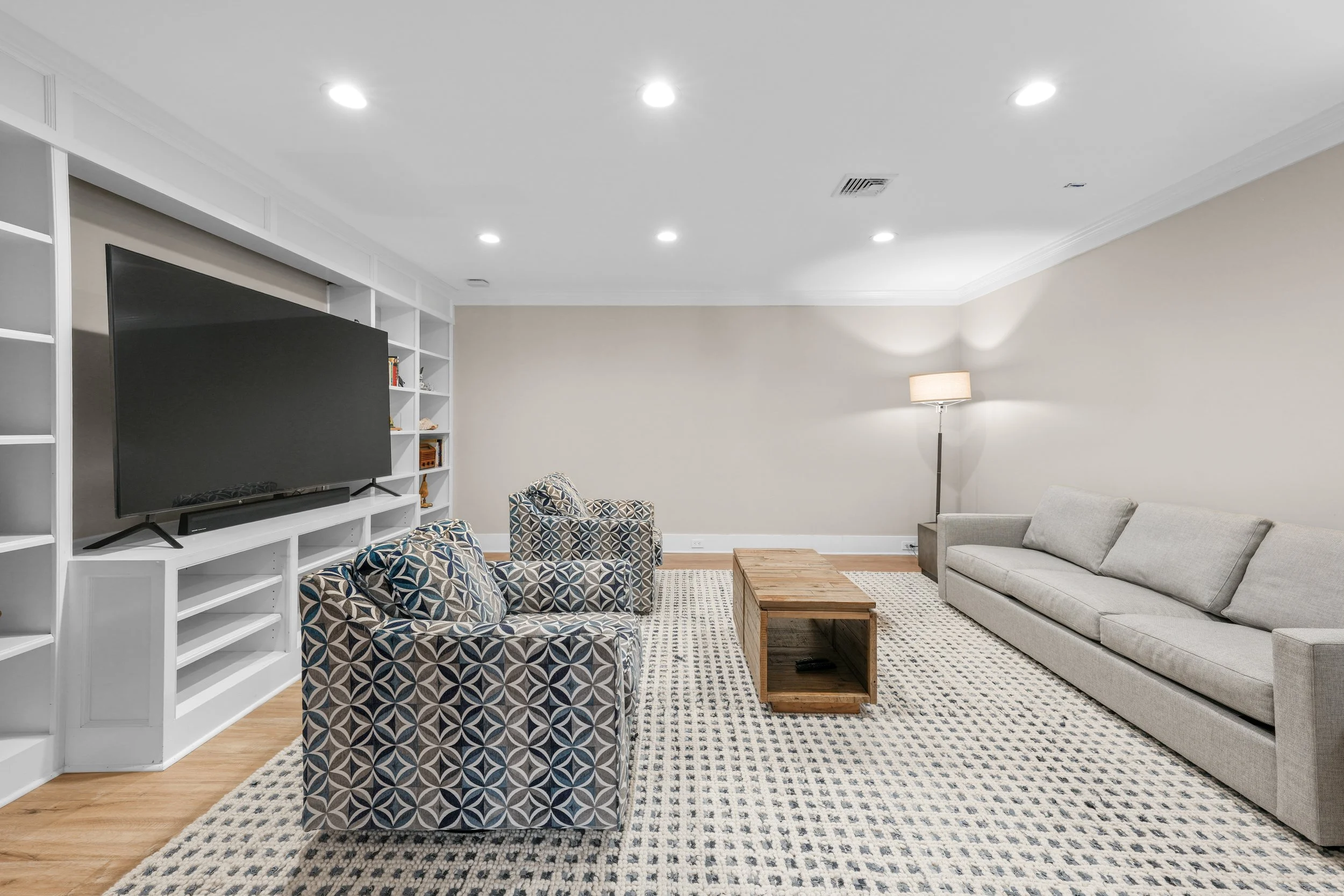 A living room with a large beige sofa, two patterned armchairs, a wooden coffee table, a floor lamp, and a wall-mounted flat-screen TV with white shelves behind it. The floor is wooden, and there is a patterned area rug.