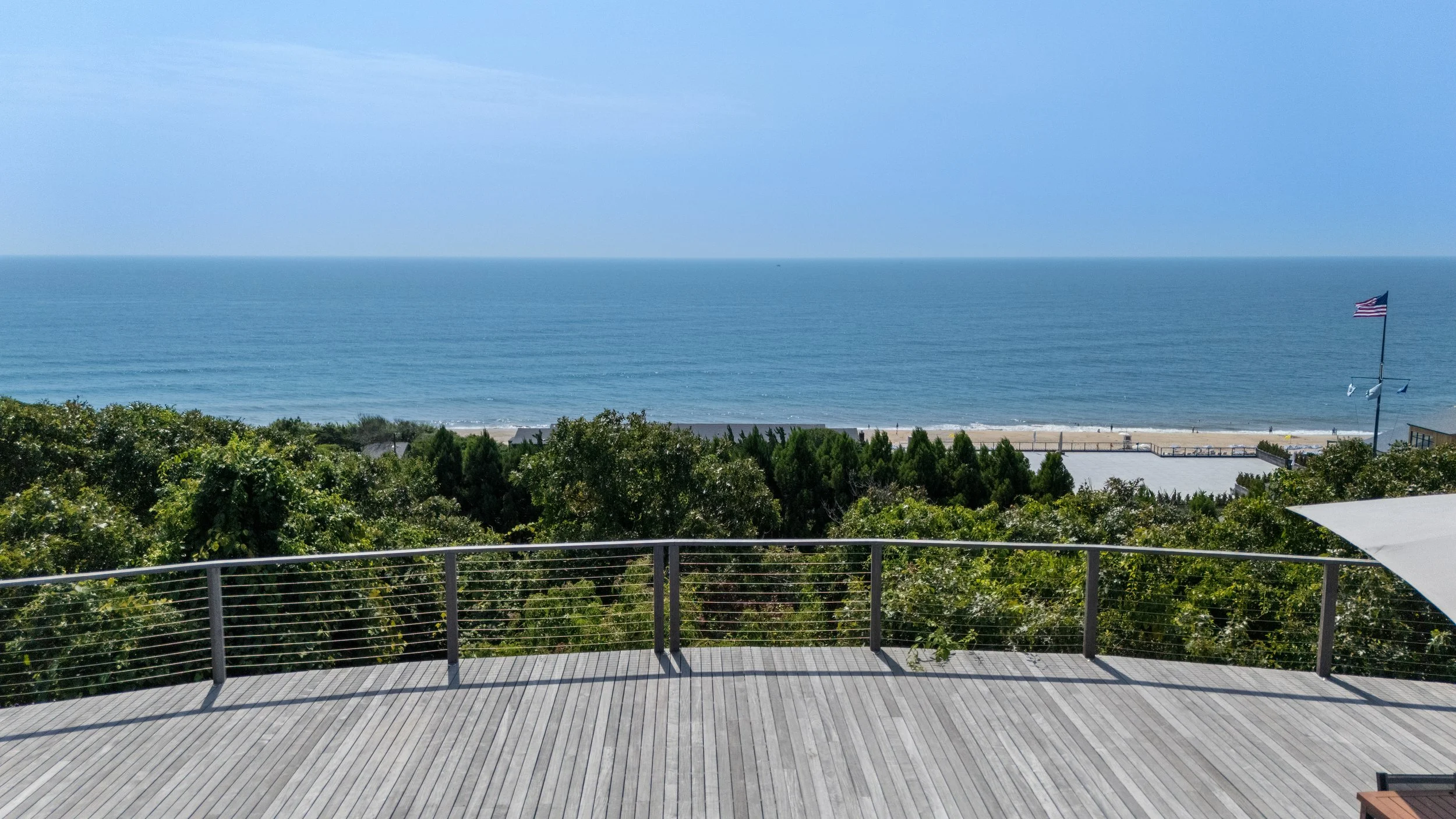 A wooden deck with a metal railing overlooks a lush green area leading to a sandy beach and the ocean in the distance, with an American flag flying on a flagpole to the right.
