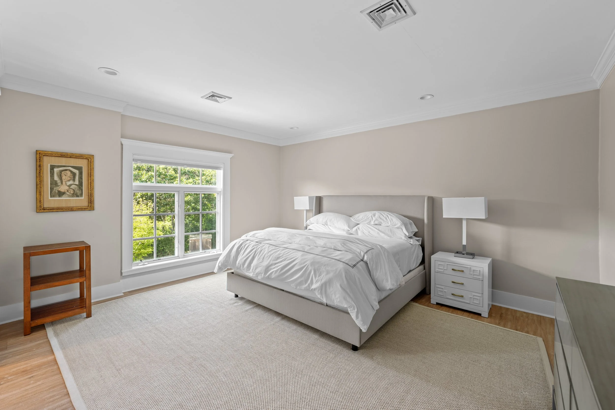 A bedroom with white walls, a large window with greenery outside, a beige bed with white bedding, two white bedside lamps, a small wooden shelf, a framed artwork on the wall, and a beige area rug on light wood flooring.