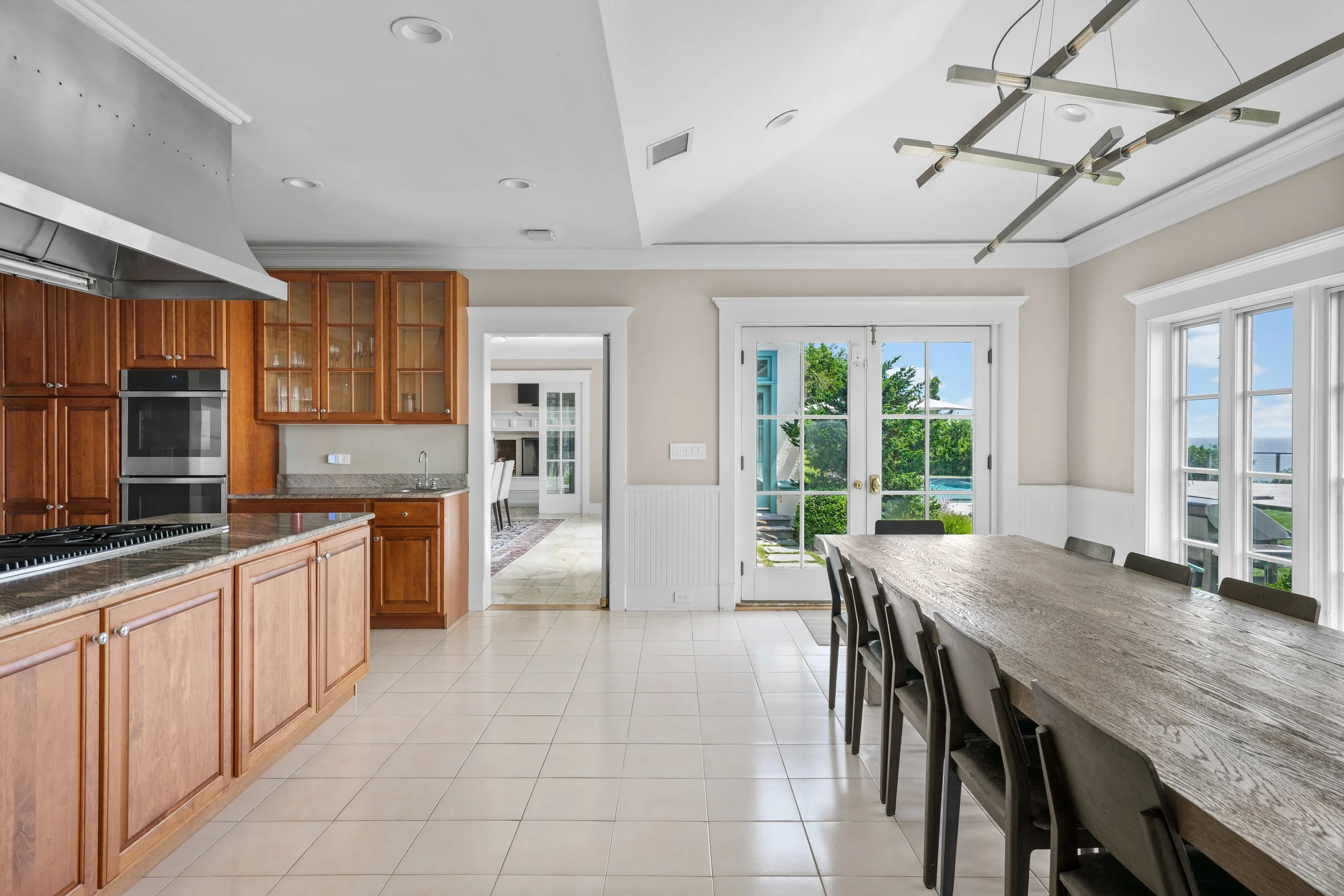 Bright kitchen and dining area with wooden cabinets, a large dining table, and glass doors leading outside with a view of greenery and sky.