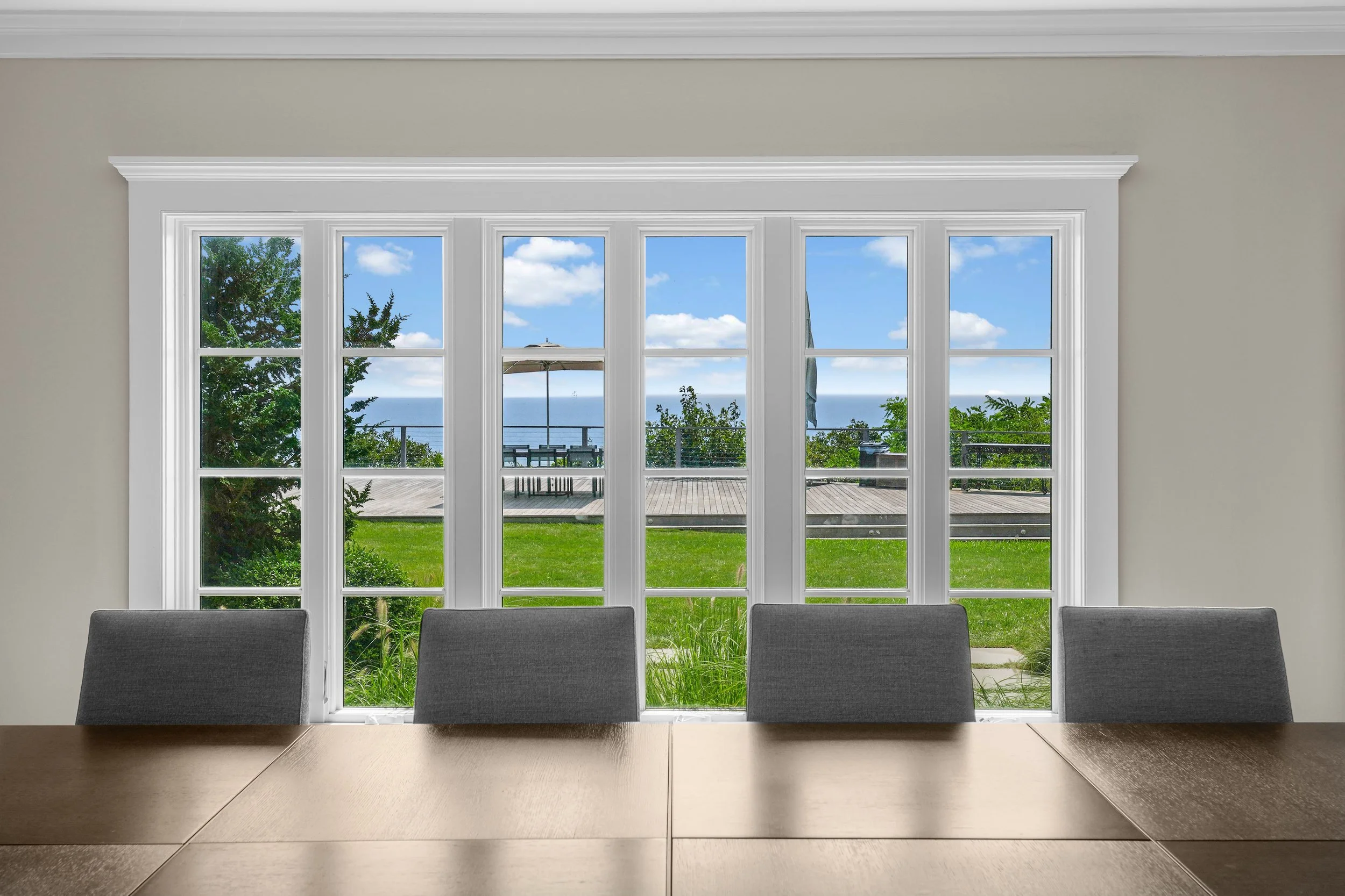 A conference room with a wooden table and four gray chairs, looking out through large white-framed windows to a lawn, patio with table and umbrella, trees, and the ocean in the distance under a blue sky with clouds.