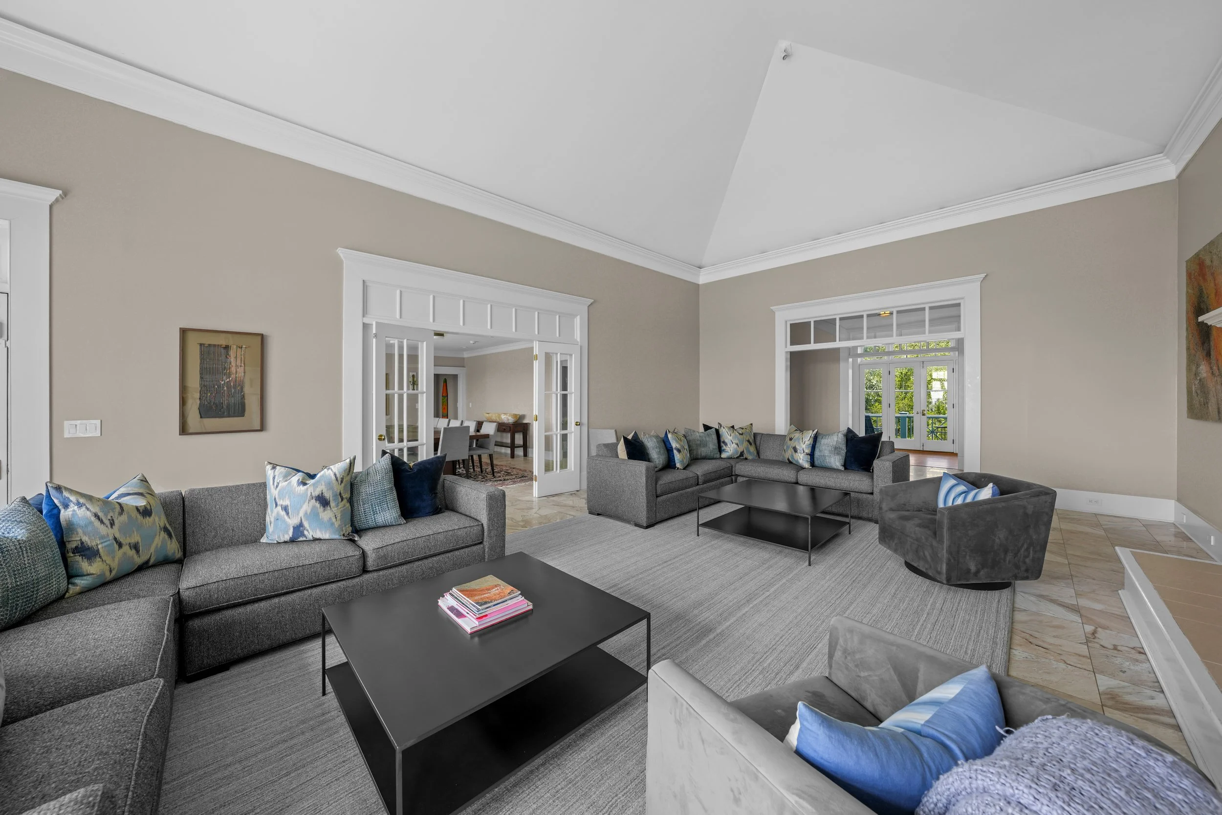 Living room with two gray sectional sofas, one gray armchair, a black coffee table, colorful pillows, a large area rug, and a view into a dining room with a table and chairs through open double doors.