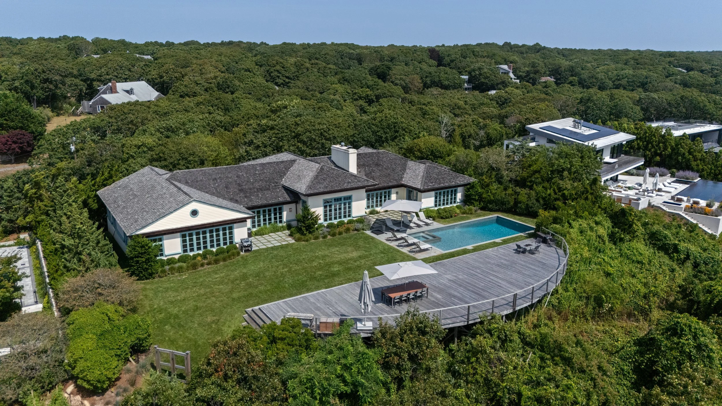 Aerial view of a modern house with a large wooden deck, a swimming pool, and lush greenery surrounding the property.