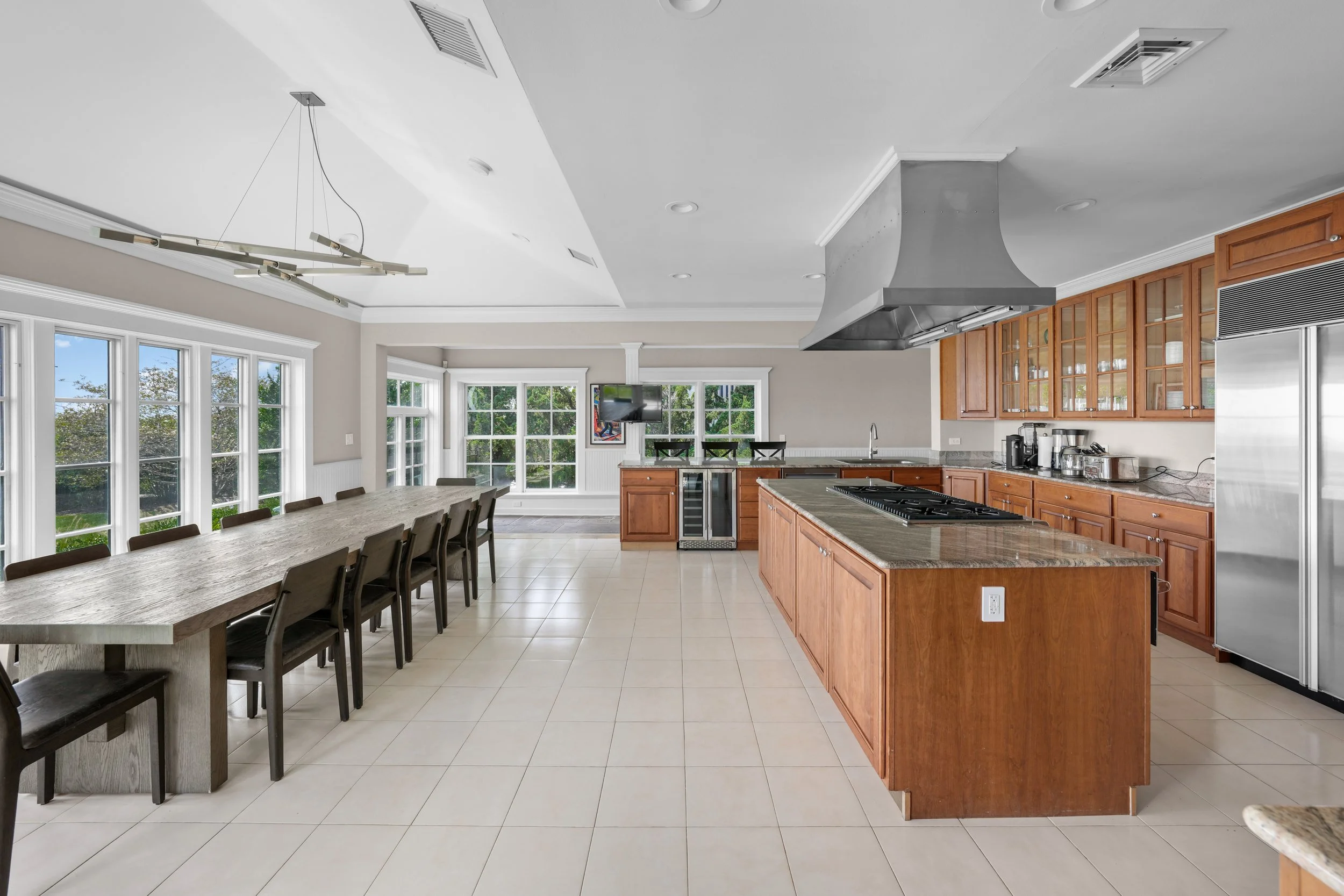 Spacious kitchen with multiple windows, wooden cabinets, island with cooktop, and a long dining table with chairs.