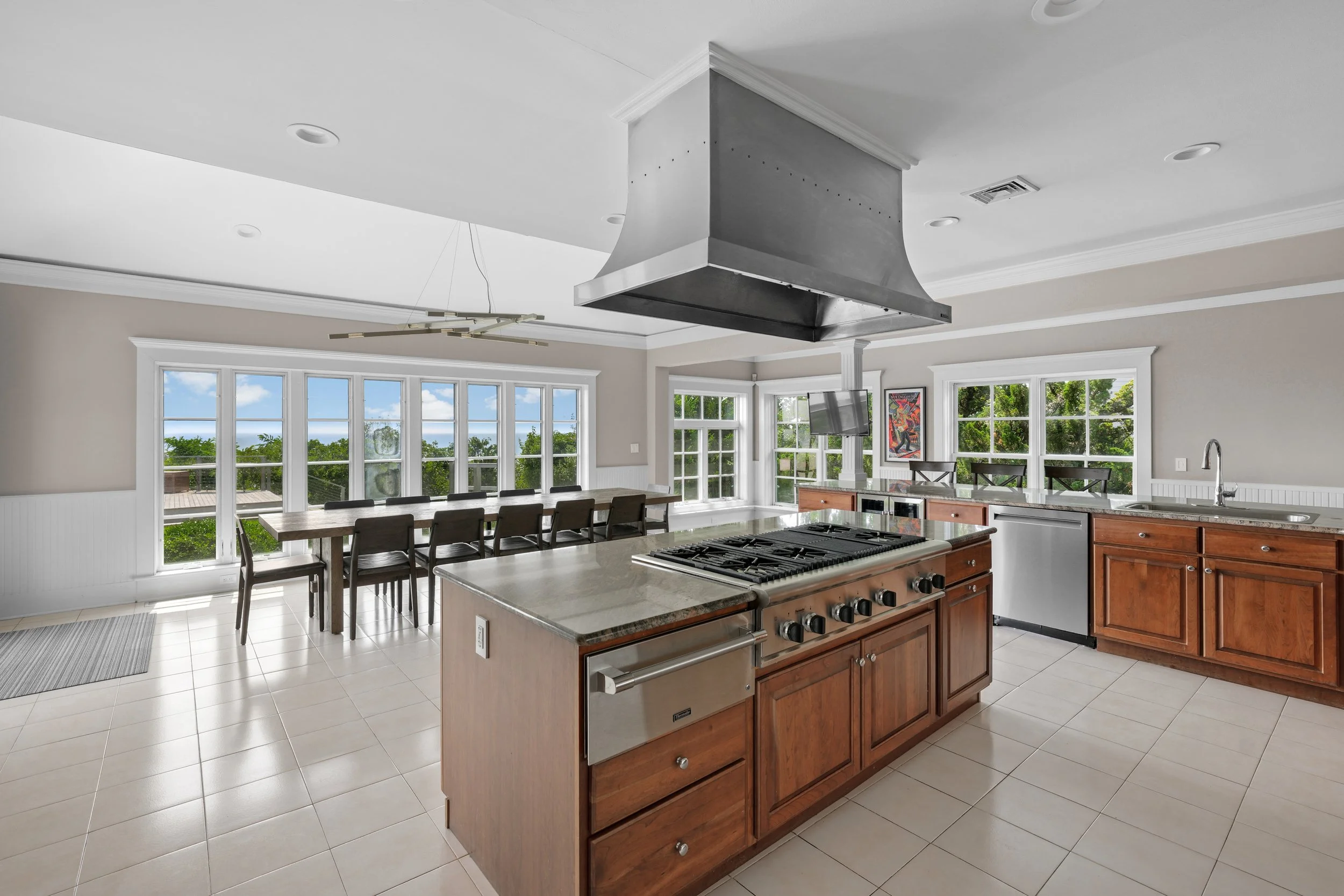 Bright kitchen with large windows, wooden cabinets, kitchen island with stove, and dining table with chairs outside.