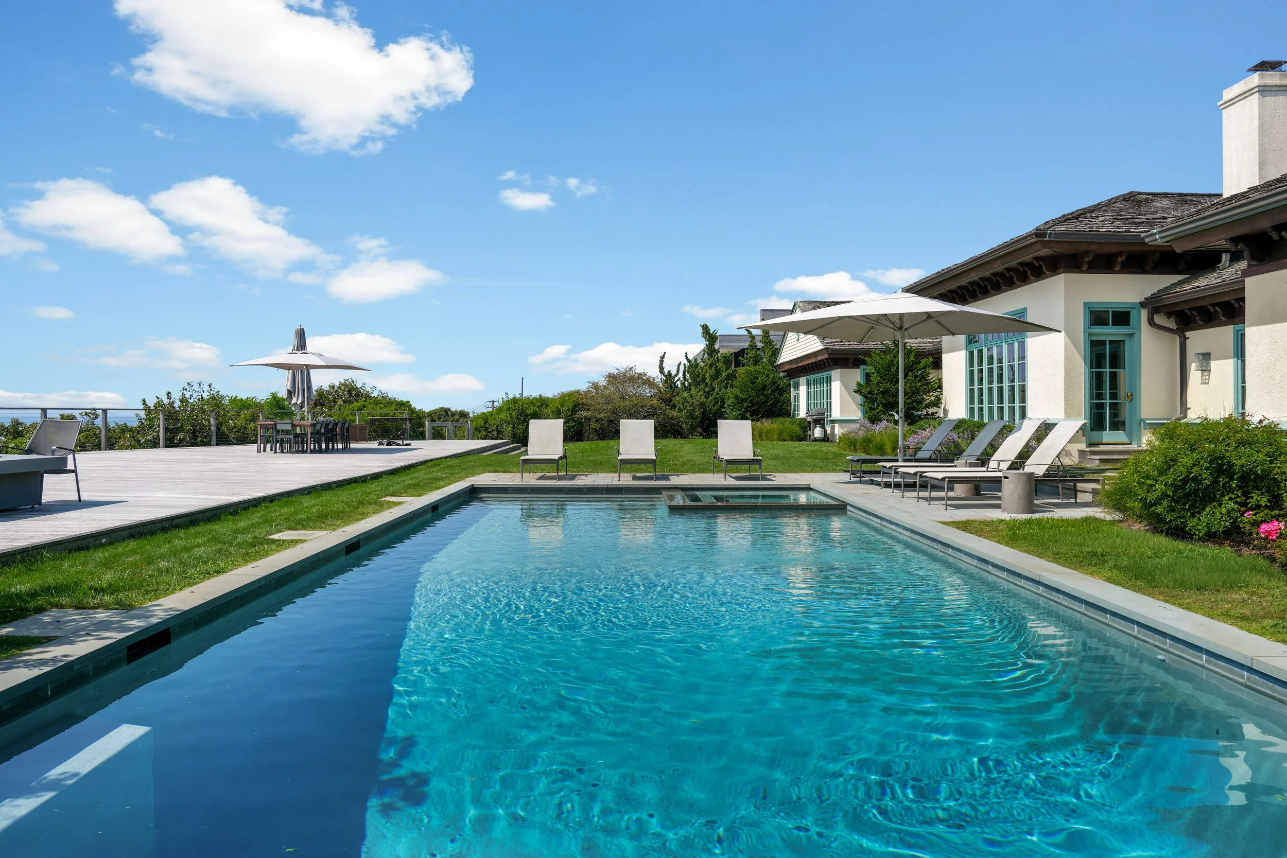 Luxury residential backyard with a rectangular swimming pool, lounge chairs with umbrellas, a wooden deck area, and a house with large windows and a sloped roof under a partly cloudy blue sky.