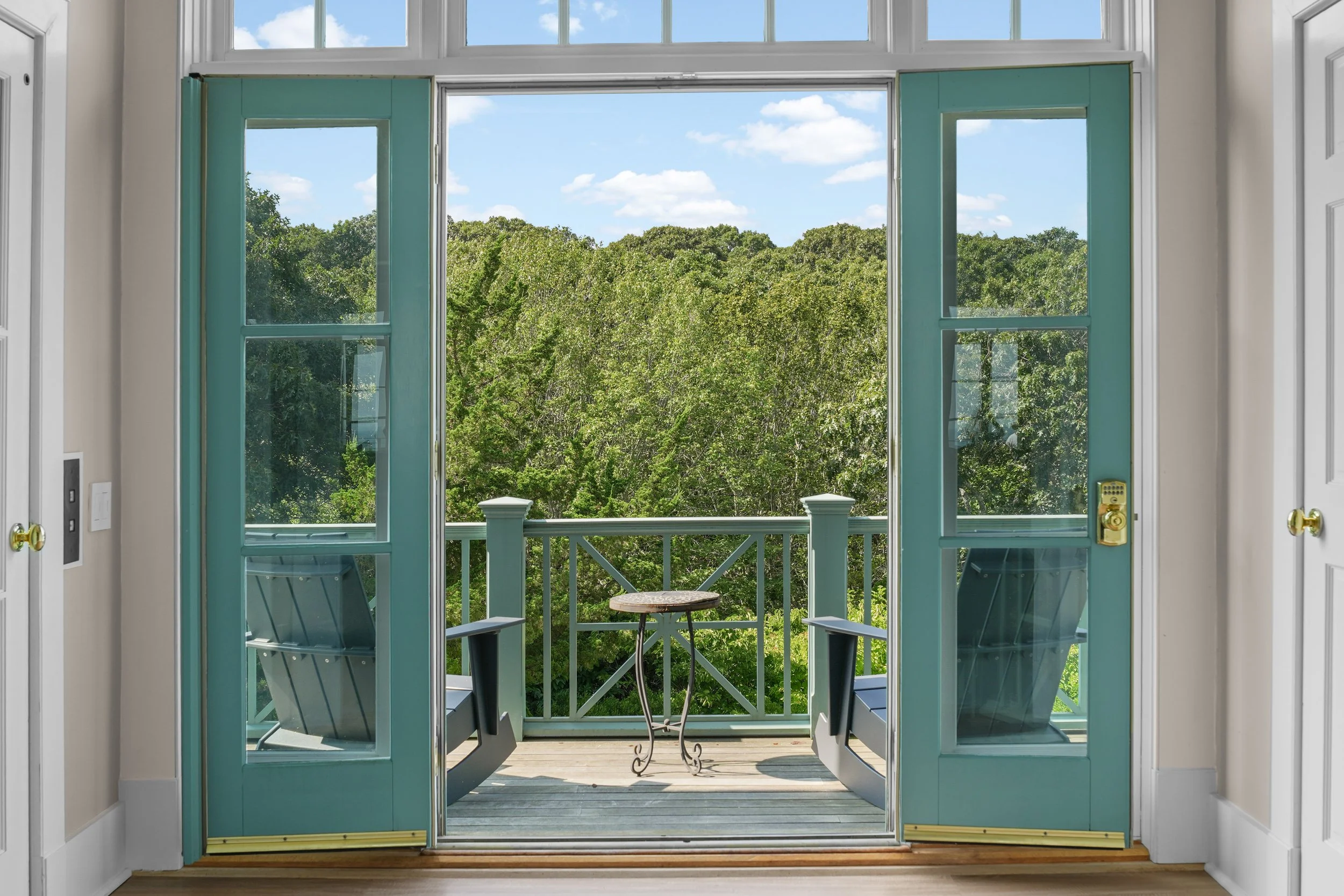 Open glass door leading to a small balcony with chairs and a small table, overlooking lush green trees and a partly cloudy blue sky.