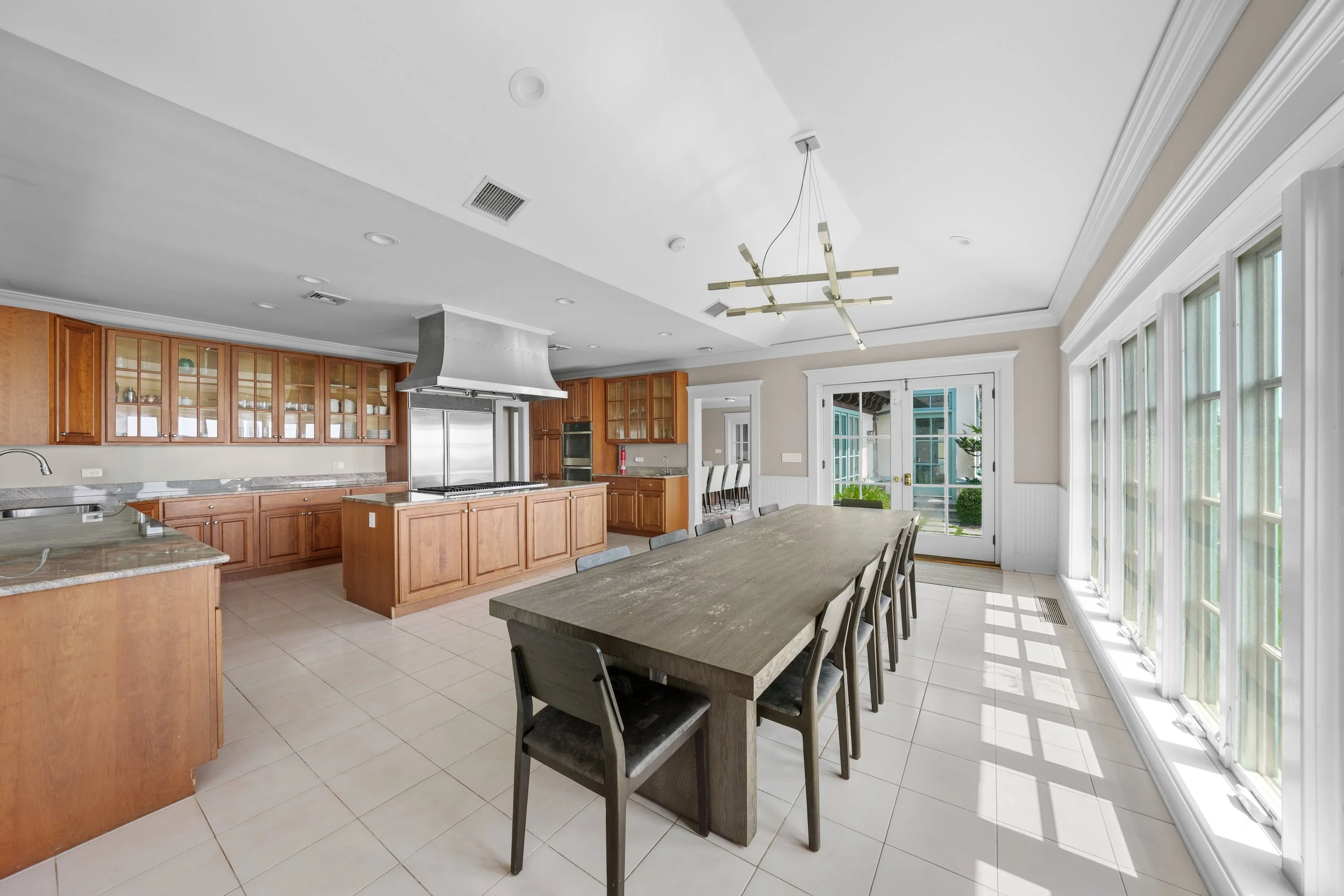 Bright kitchen with wood cabinets, a large island, a stainless steel range hood, and a dining table with chairs near large windows and glass doors leading outside.