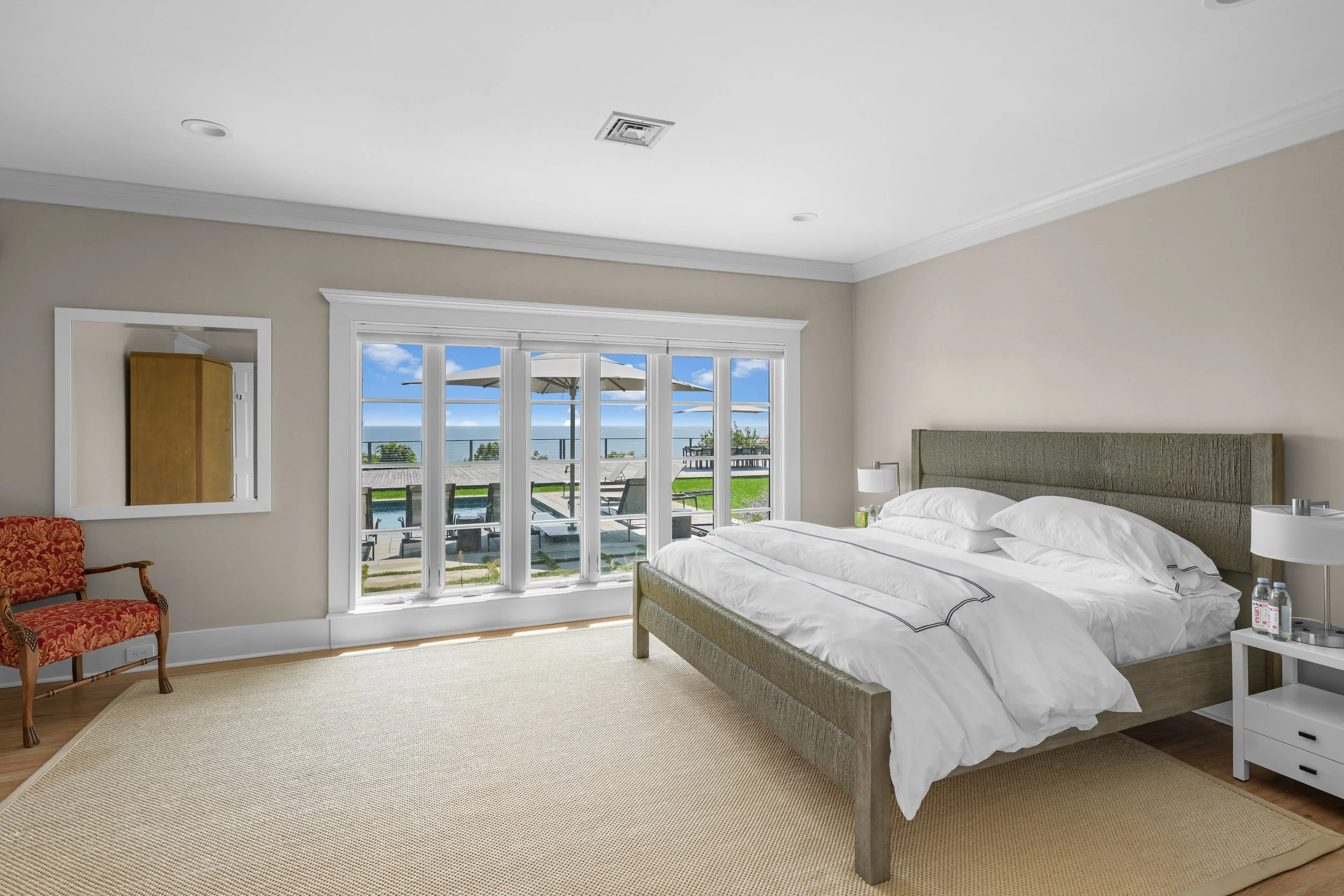 Bright hotel bedroom with large window view of outdoor pool, lounge chairs, and umbrellas, featuring a bed with a green upholstered headboard, white bedding, bedside tables, lamps, and a decorative chair.