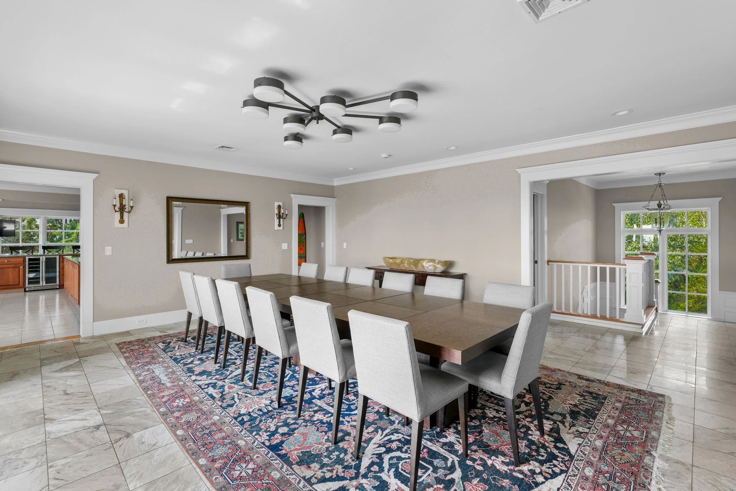 A spacious dining room with a long wooden table surrounded by fifteen light gray upholstered chairs, a decorative rug underneath, and a modern black chandelier overhead. There's a large mirror on the beige wall and a sideboard with a decorative bowl.