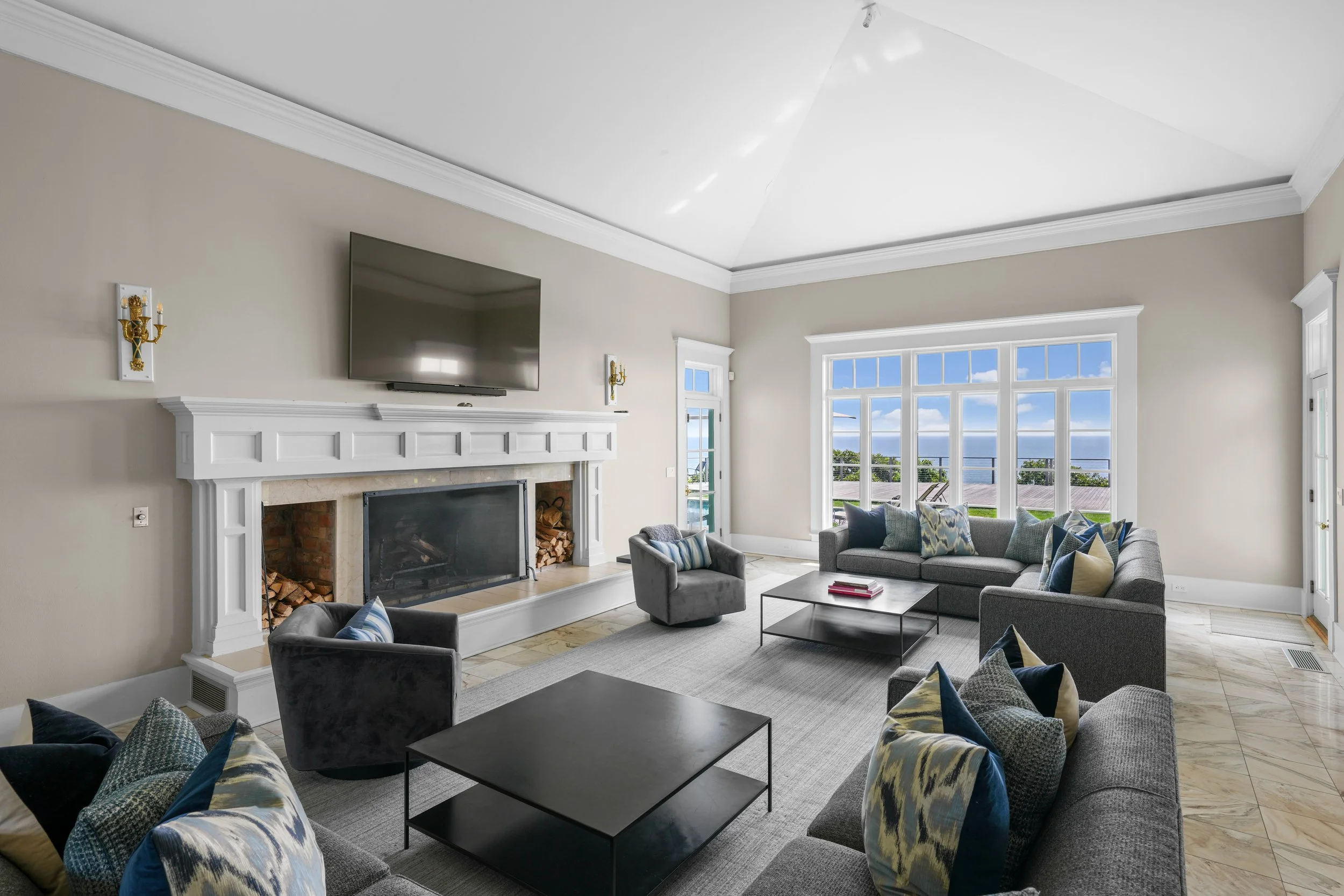 Living room with large windows showing ocean view, gray sofas with colorful pillows, a fireplace, wall-mounted TV, and coffee tables.