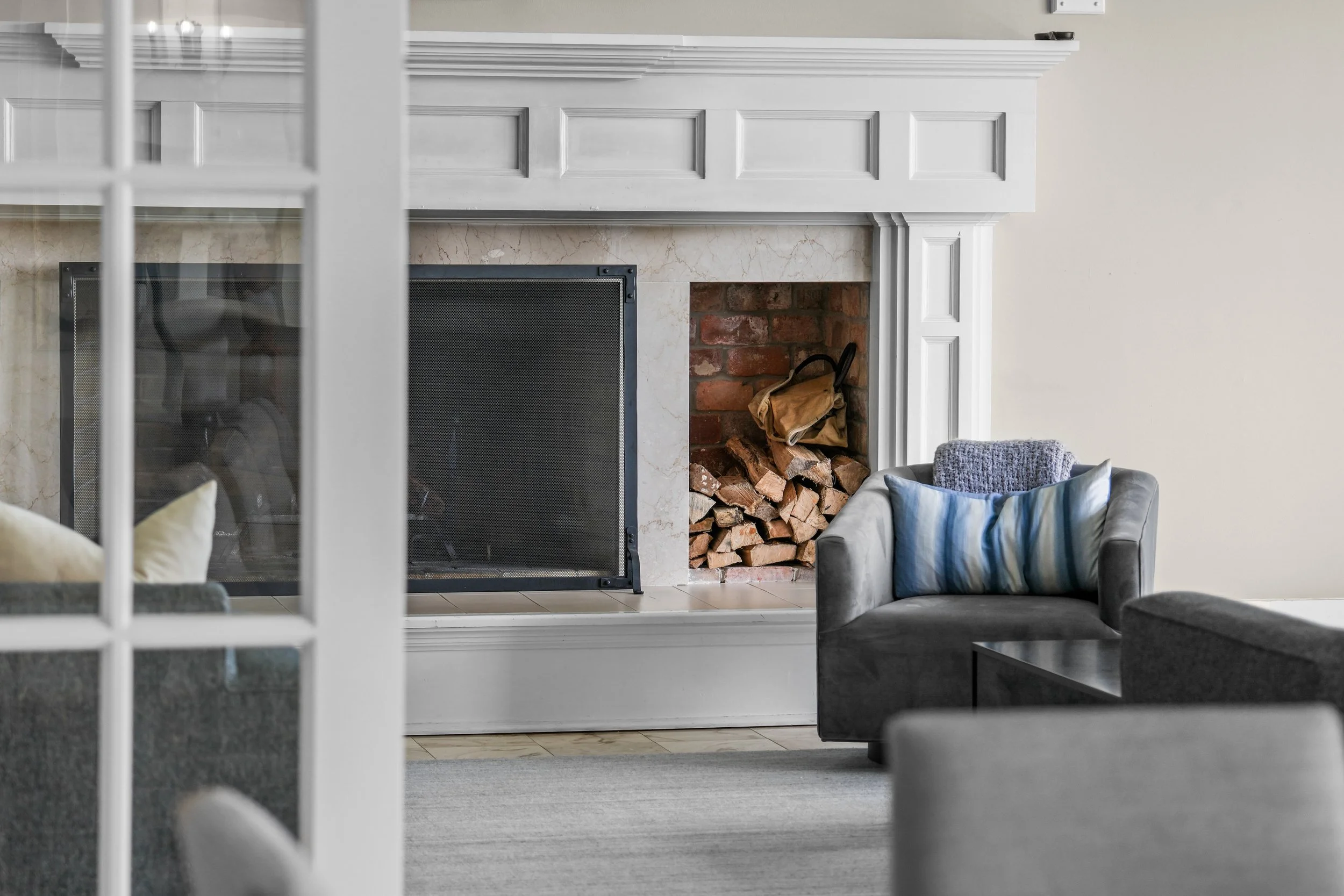 Living room with a white fireplace, logs stored inside, a gray armchair with a blue pillow, a blue throw blanket, and a gray sofa with a pillow. A sliding glass door partially visible on the left.
