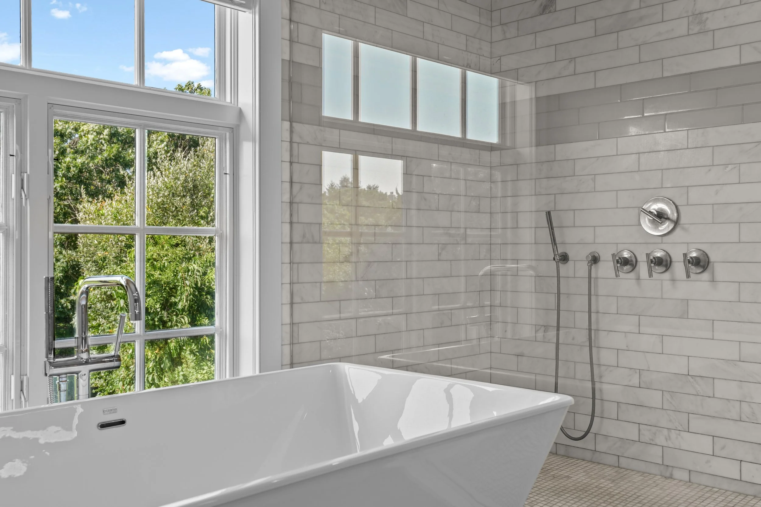 Modern bathroom with a white bathtub next to large window with a view of green trees, white tiled walls, and a shower area with multiple controls and a handheld showerhead.