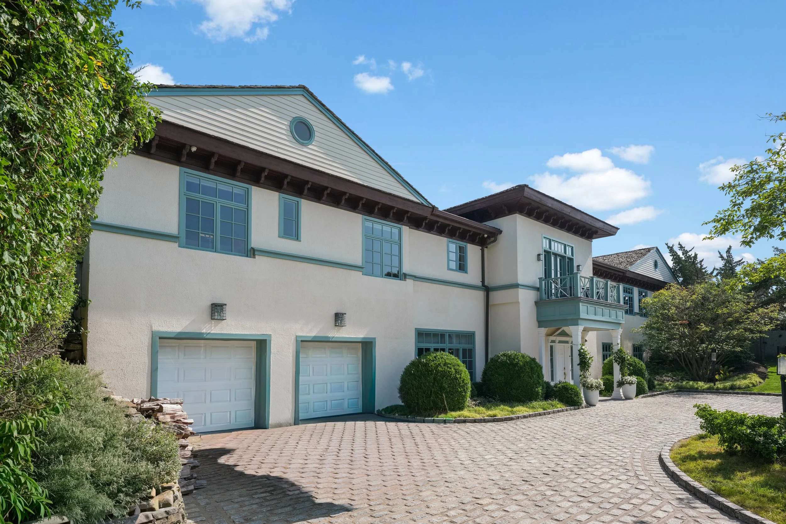 A large, modern house with white walls and blue accents, a cobblestone driveway, and lush greenery under a partly cloudy blue sky.