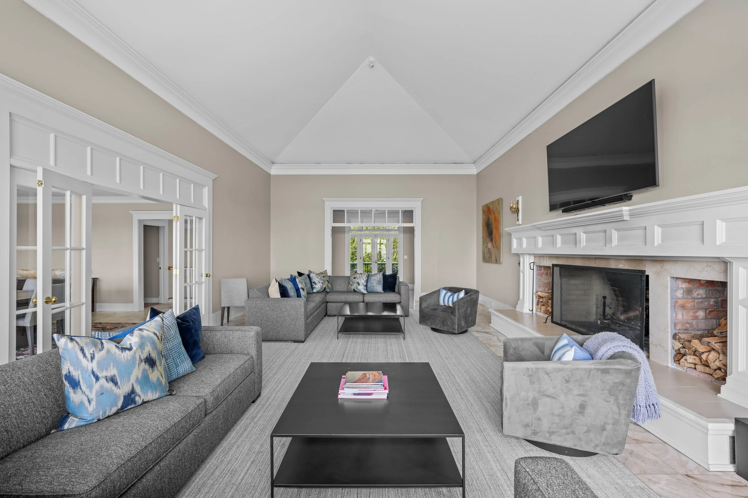Bright living room with beige walls, white crown molding, a brick fireplace with a mounted TV, gray sofas and armchairs with blue and patterned pillows, black coffee table, and large window in the background.