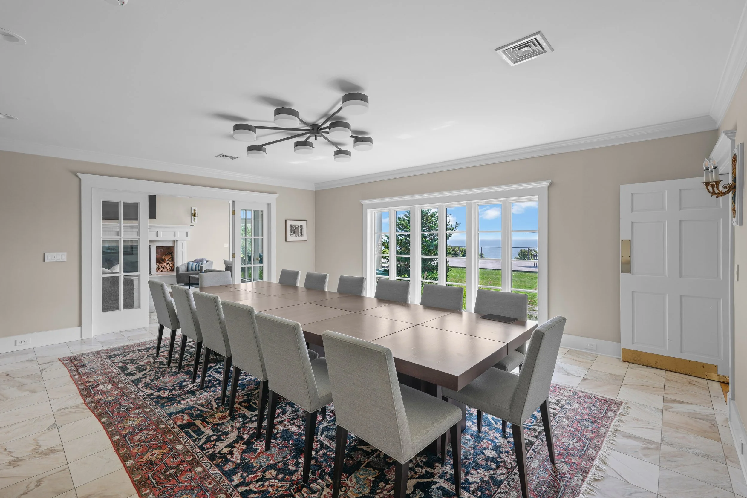 Bright dining room with a large wooden table, 12 gray upholstered chairs, large windows showing a green outdoor space, traditional fireplace in the adjacent living room, and a patterned area rug on tiled floor.