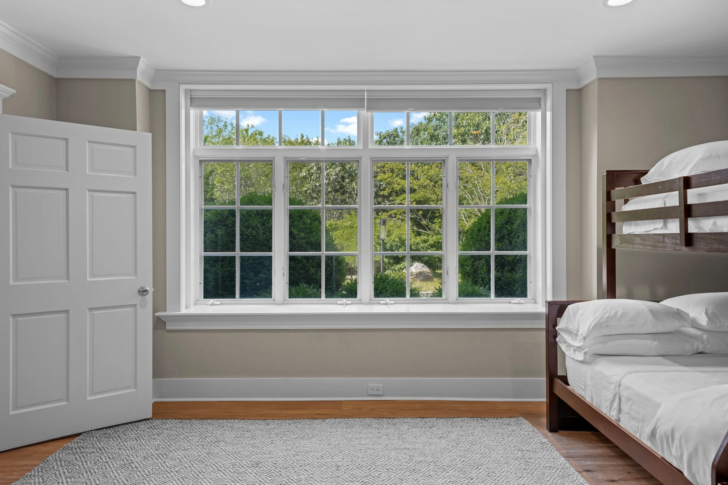 Empty bedroom with large window, bed with wooden frame, beige walls, hardwood floor, and gray rug.