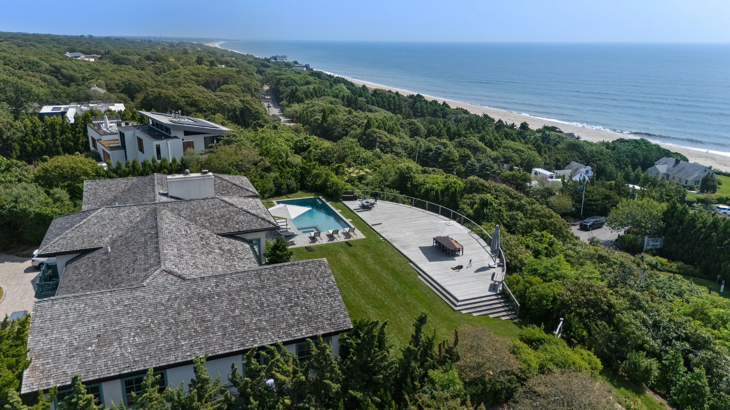 Aerial view of a coastal property with a swimming pool, lounge area, and a large deck, surrounded by lush green trees and overlooking the ocean and beach.