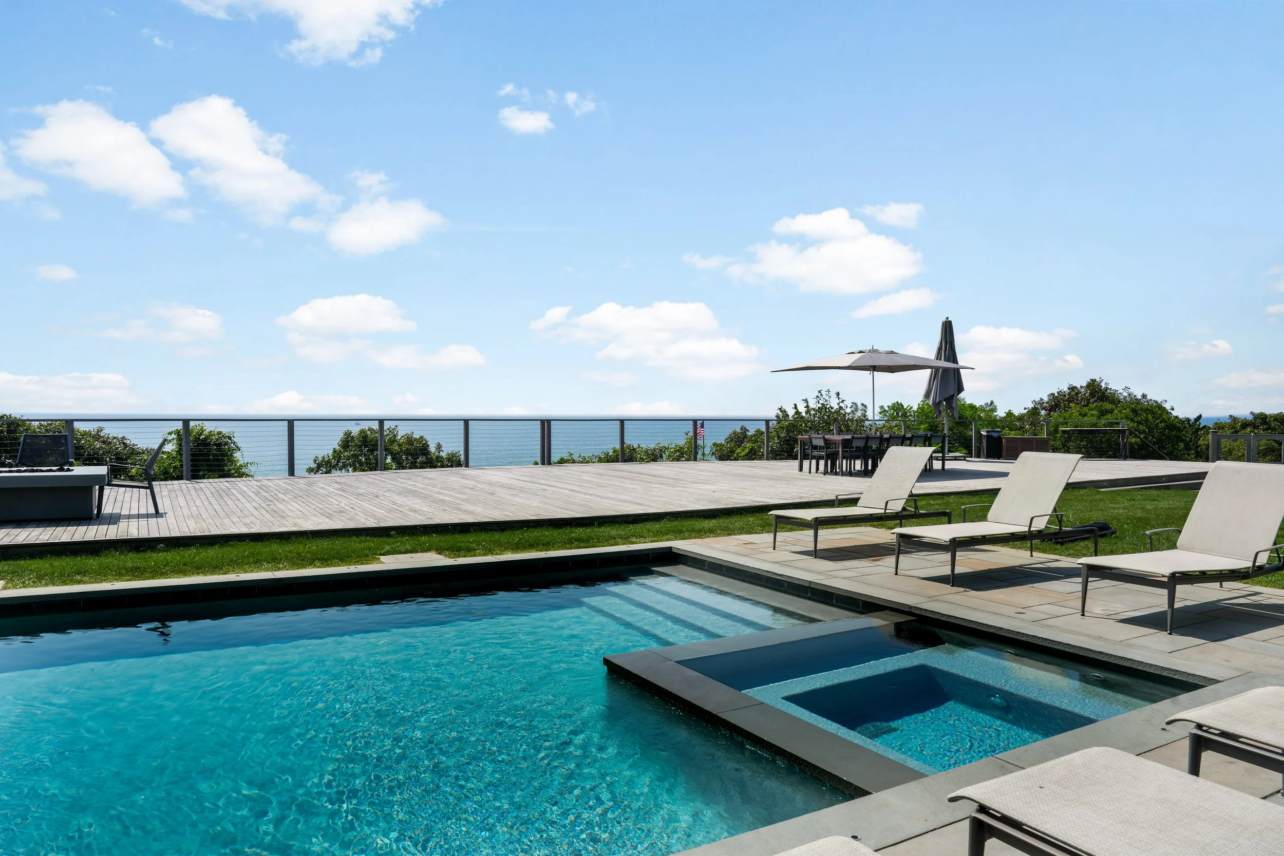Outdoor swimming pool with a hot tub, lounge chairs, a wooden deck, a table with an umbrella, and a view of the ocean.
