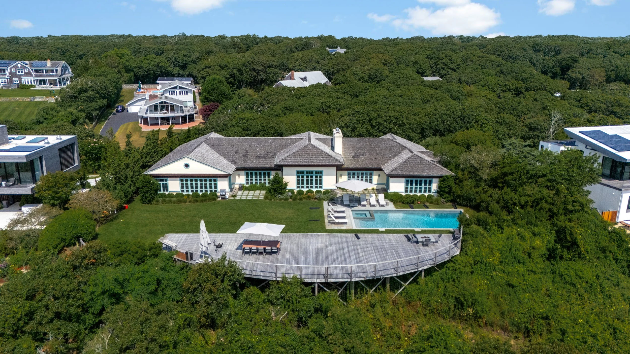 A large house with beige walls, a gray roof, and multiple patio doors, surrounded by greenery. In the backyard, there is a wooden deck with outdoor furniture, a rectangular swimming pool, and lounge chairs with umbrellas. The property is situated on